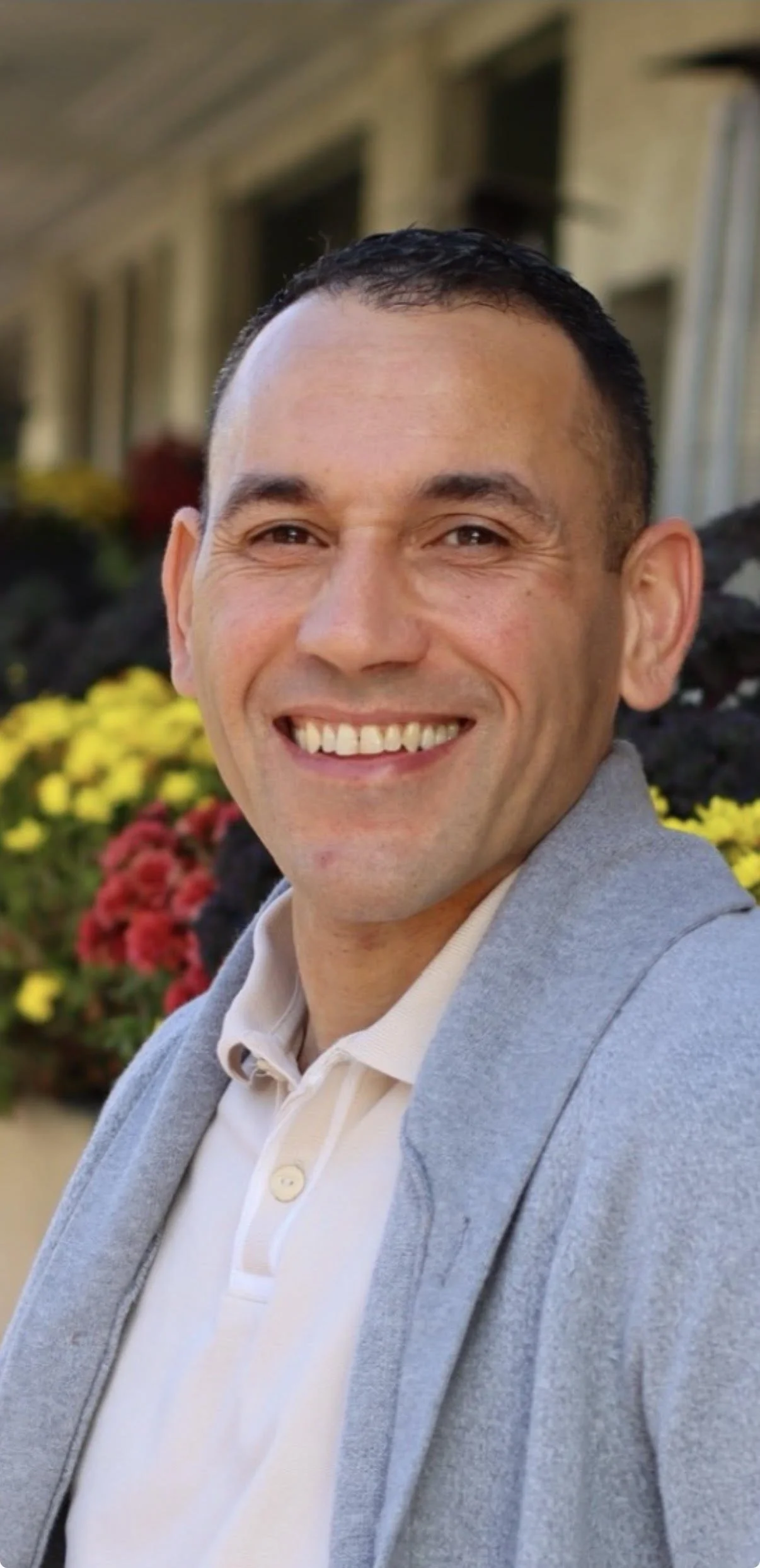 A smiling man with short dark hair, wearing a light gray blazer over a beige polo shirt, standing outdoors with a background of yellow and red flowers and a building.