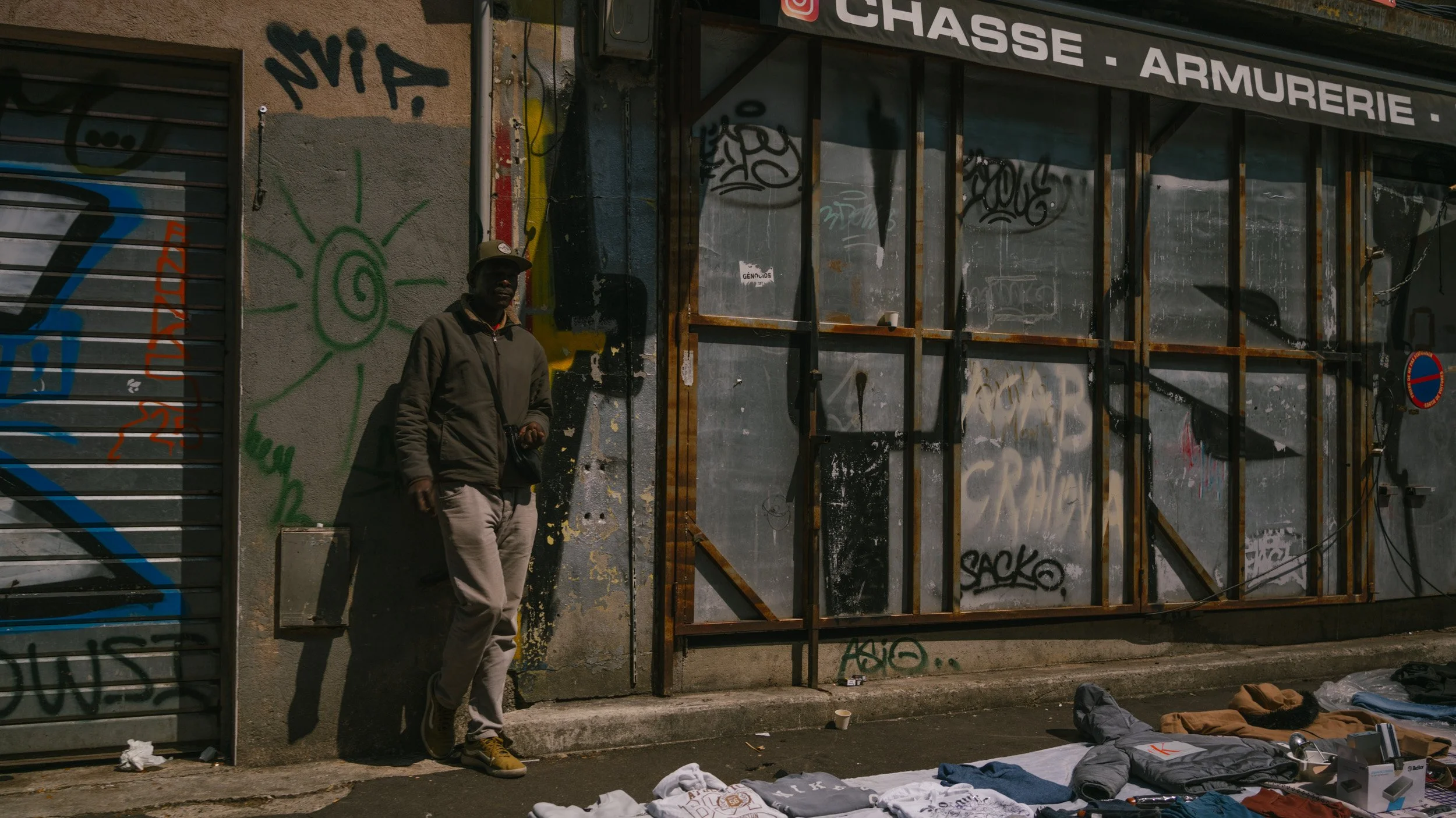 Ein Mann steht vor einer Wand mit Graffiti, auf der Straße liegen Kleidung und andere Gegenstände ausgebreitet.