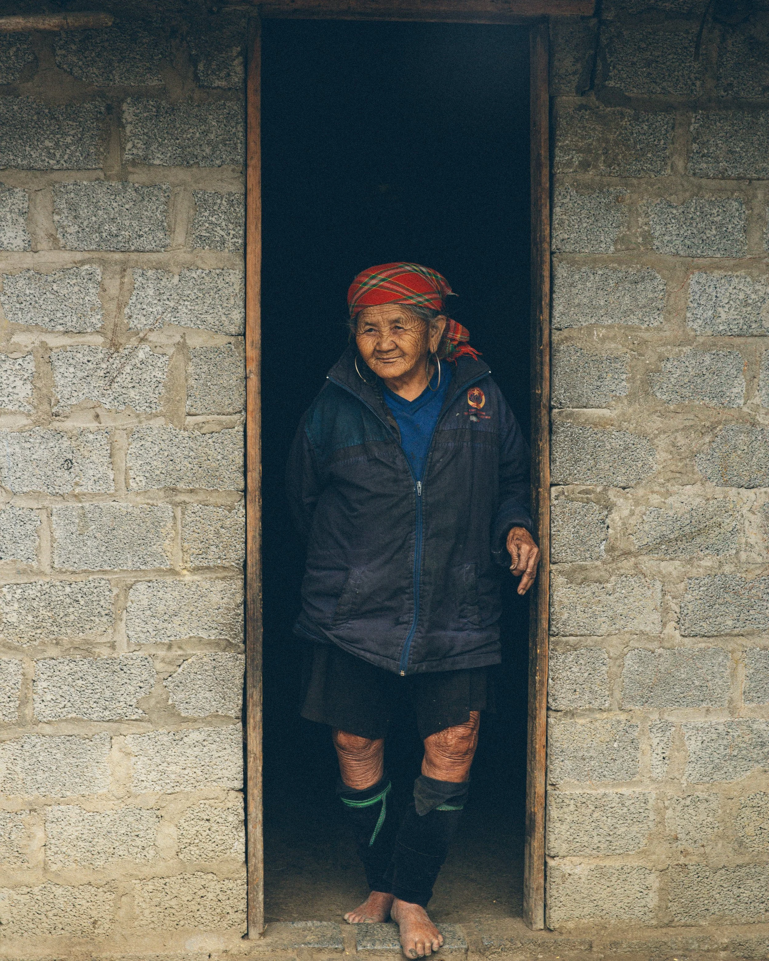 Ältere Frau im traditionellen Kleidung steht in einem kleinen Fensterrahmen einer Ziegelmauer in einem kleinen Raum, schaut nach draußen, trägt einen roten Mund- oder Kopftuch, trägt Ohrringe und Laufen barfuß.