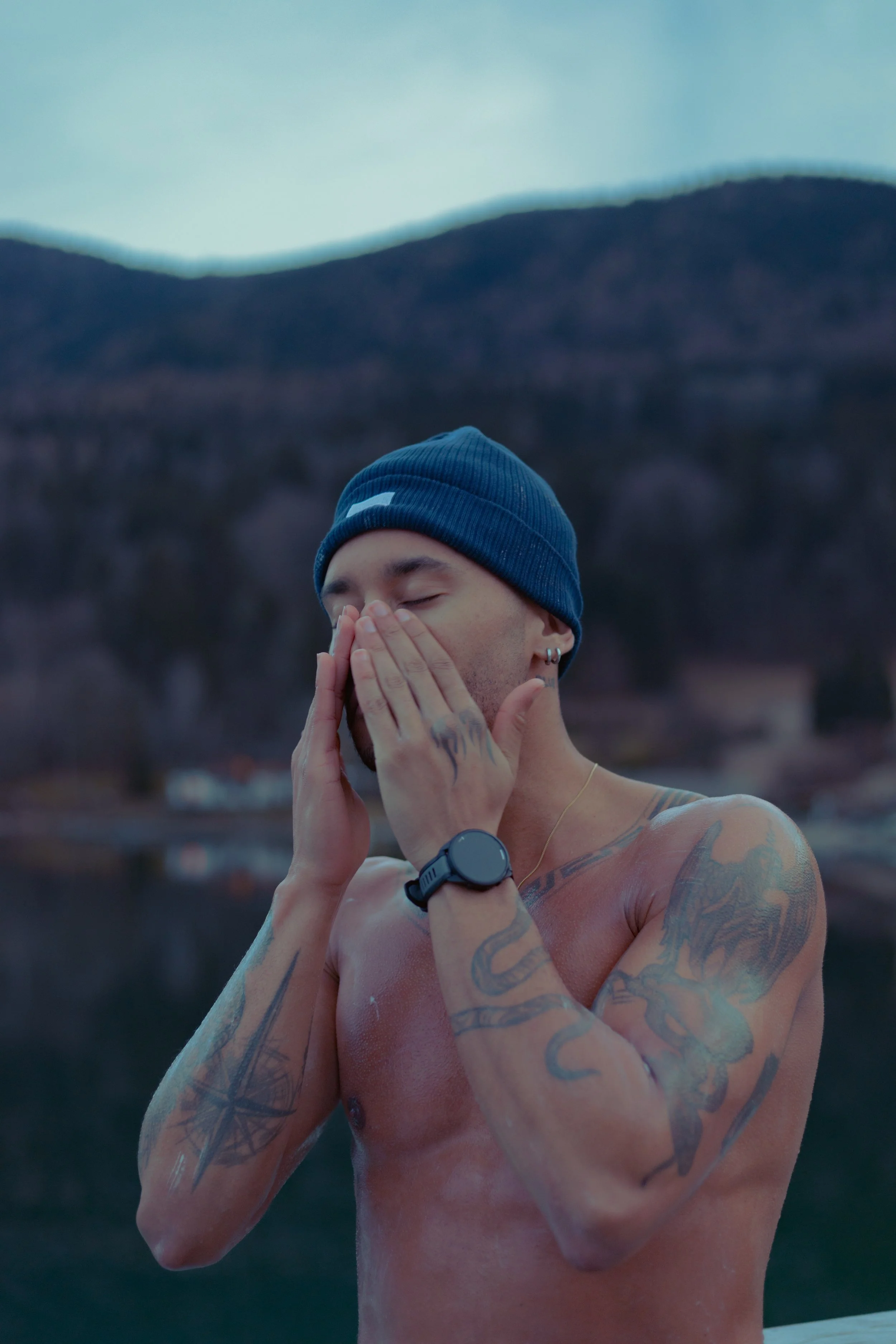 Ein nackter Mann mit Tattoos, blauer Mütze, Uhren und Piercings steht vor einer Berglandschaft und bedeckt sein Gesicht mit den Händen.