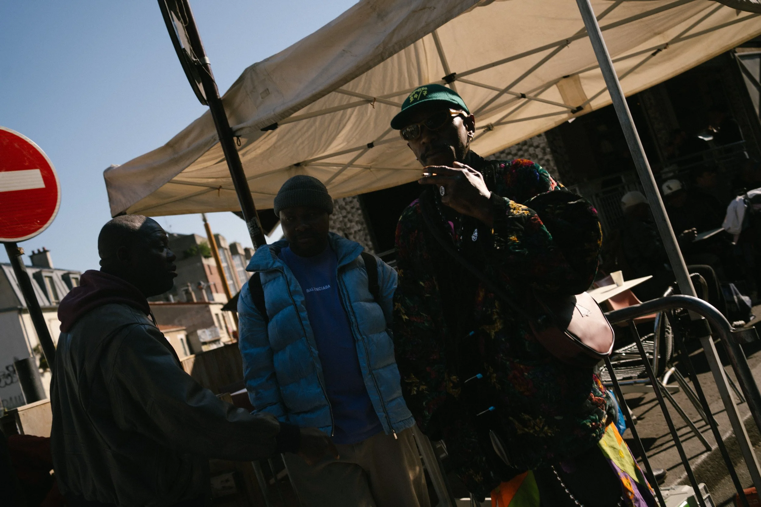 Drei Menschen stehen bei einem Marktstand im Freien, mit einem weißen Zelt und Gebäuden im Hintergrund, an einem sonnigen Tag.