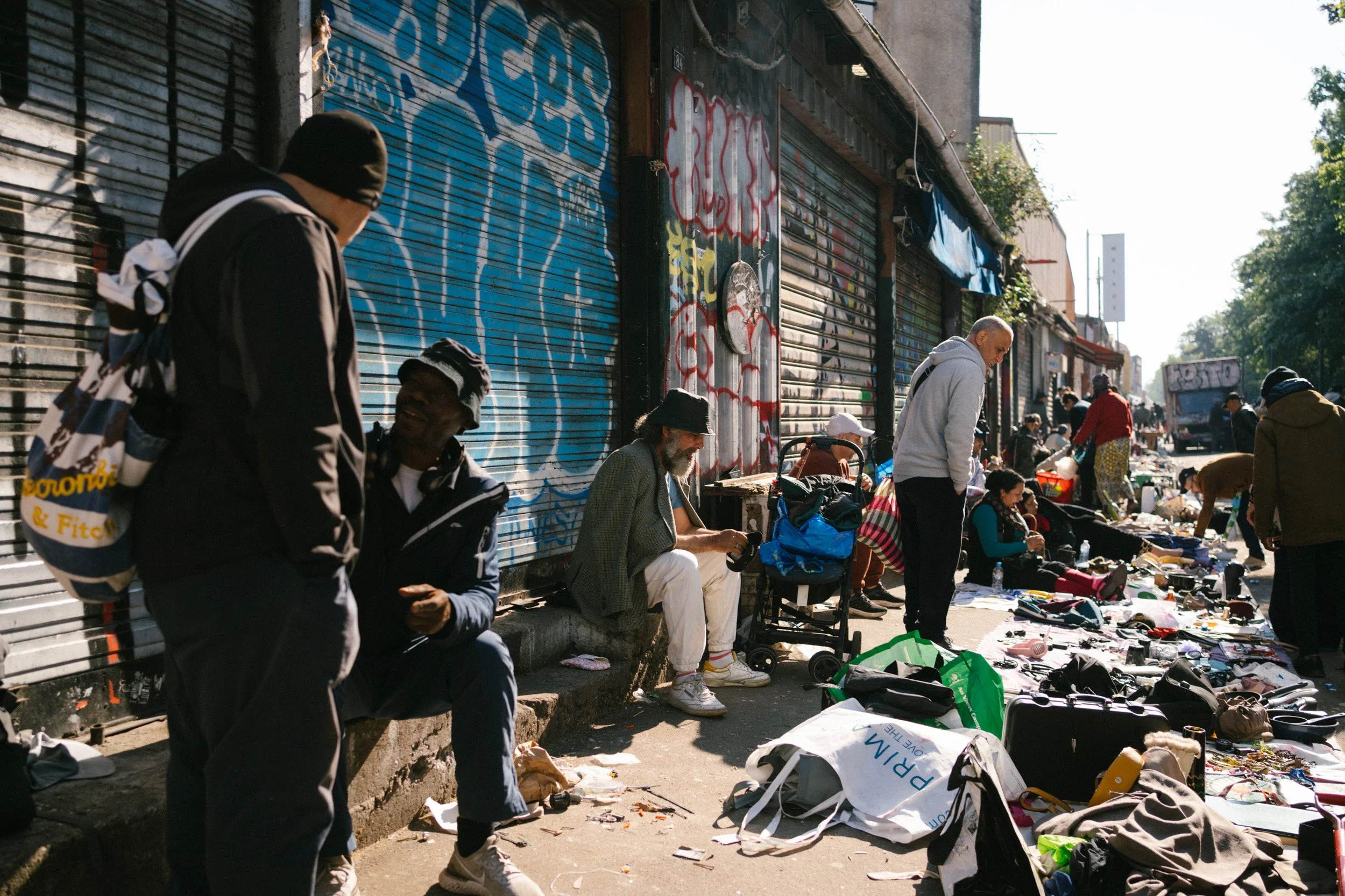 Straßenflohmarkt mit verschiedenen Menschen, die Kleidung und Gegenstände auf Tischen verkaufen, vor grauen Kodama rodeln mit Graffiti besprüht.