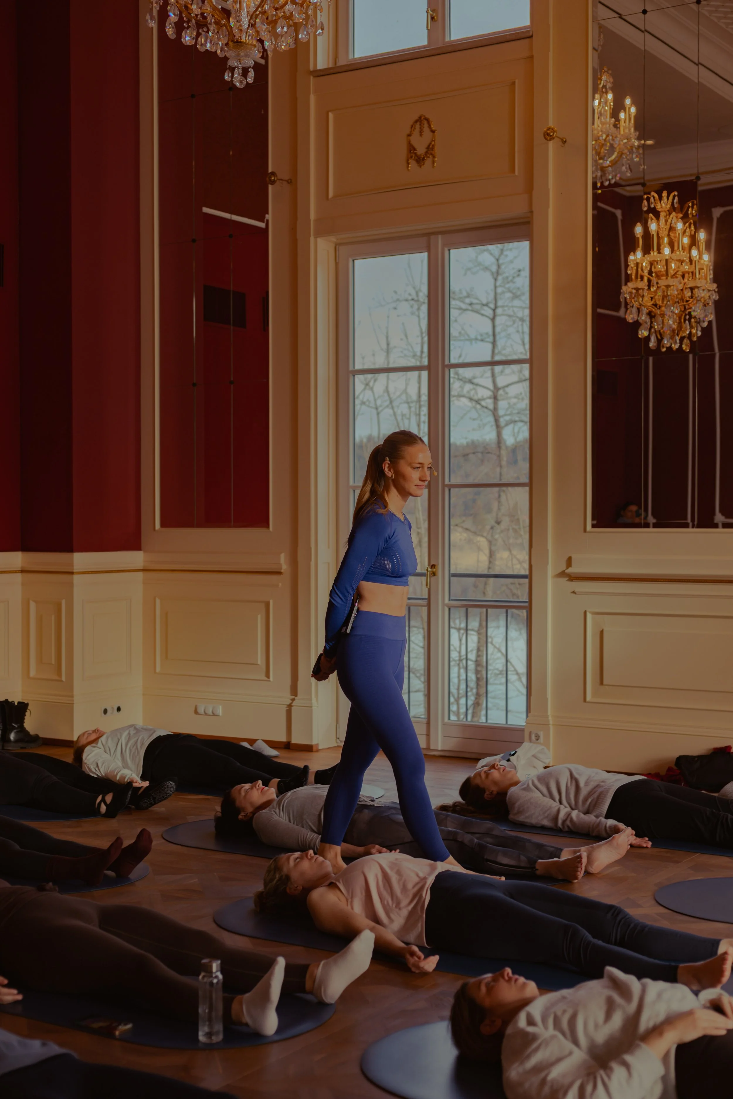 Eine Gruppe von Menschen nimmt an einem Yoga- oder Entspannungskurs in einem eleganten Raum mit hohen Fenstern und Kronleuchtern teil. Alle Personen liegen auf Yogamatten auf dem Boden, während eine Frau in blauer Sportkleidung in der Mitte steht und
