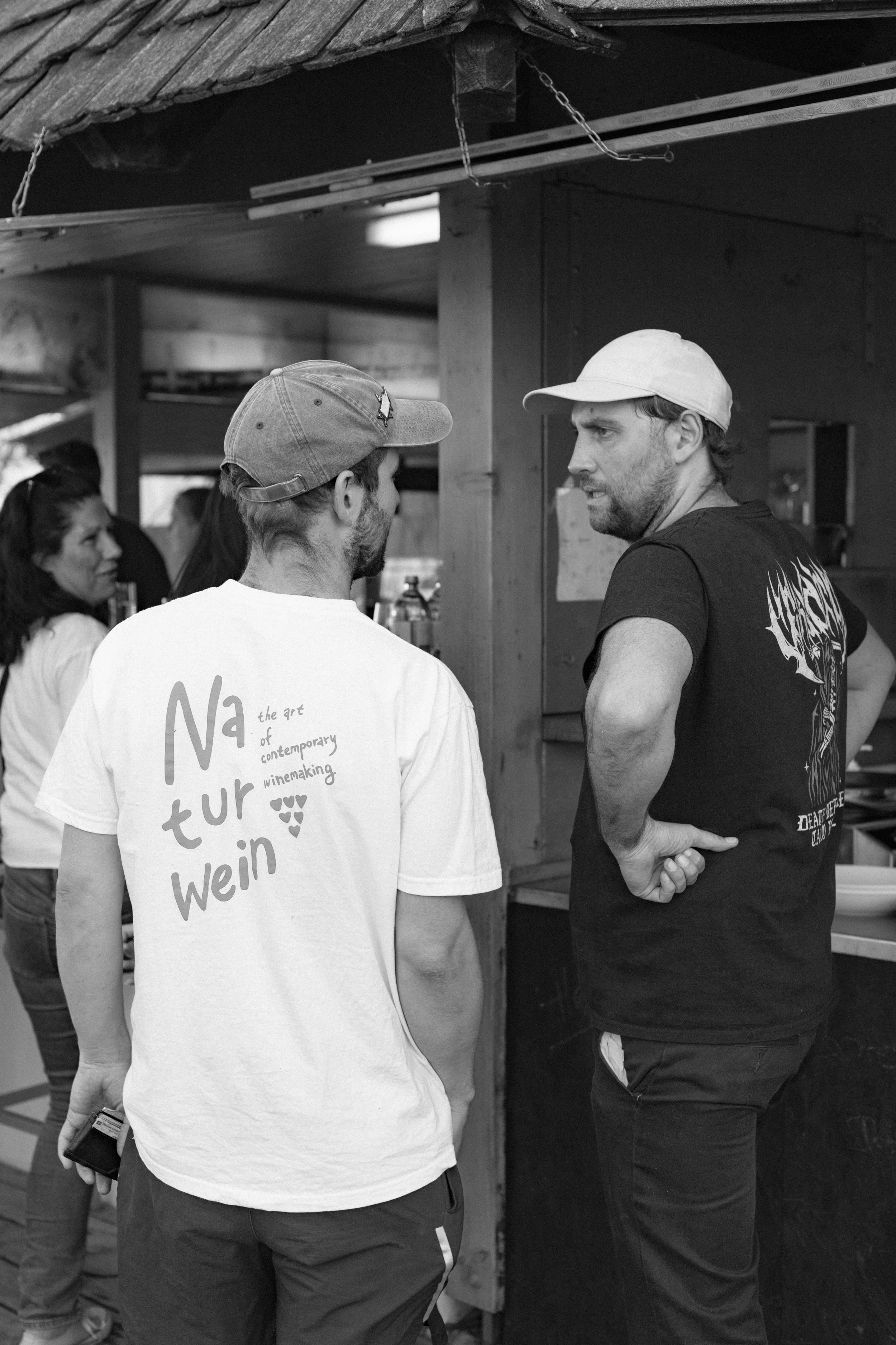 Zwei Männer unterhalten sich. Der Mann auf der linken trägt ein weißes T-Shirt mit Aufdruck, der Mann auf der rechten trägt ein schwarzes T-Shirt mit Grafik. Im Hintergrund sind weitere Menschen zu sehen.