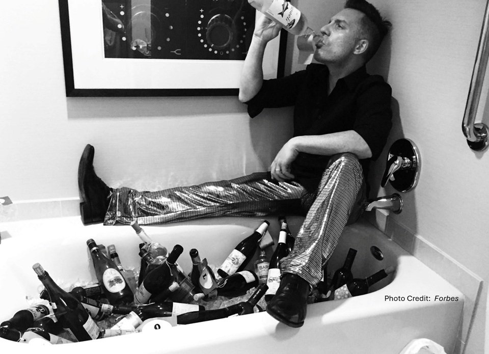 Image of a man drinking wine next to a bathtub full of wine bottles