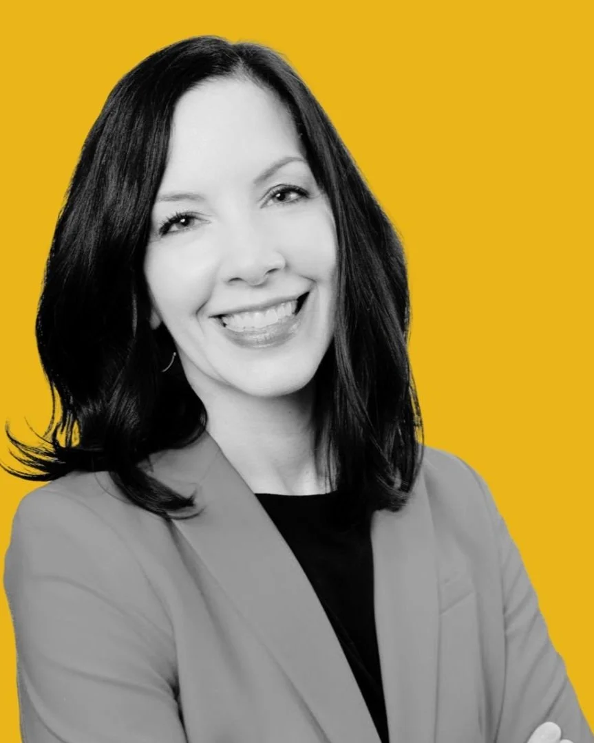 Black and white portrait of a executive coach, Angela Justice, PhD with shoulder-length dark hair, wearing professional attire, against a yellow background.