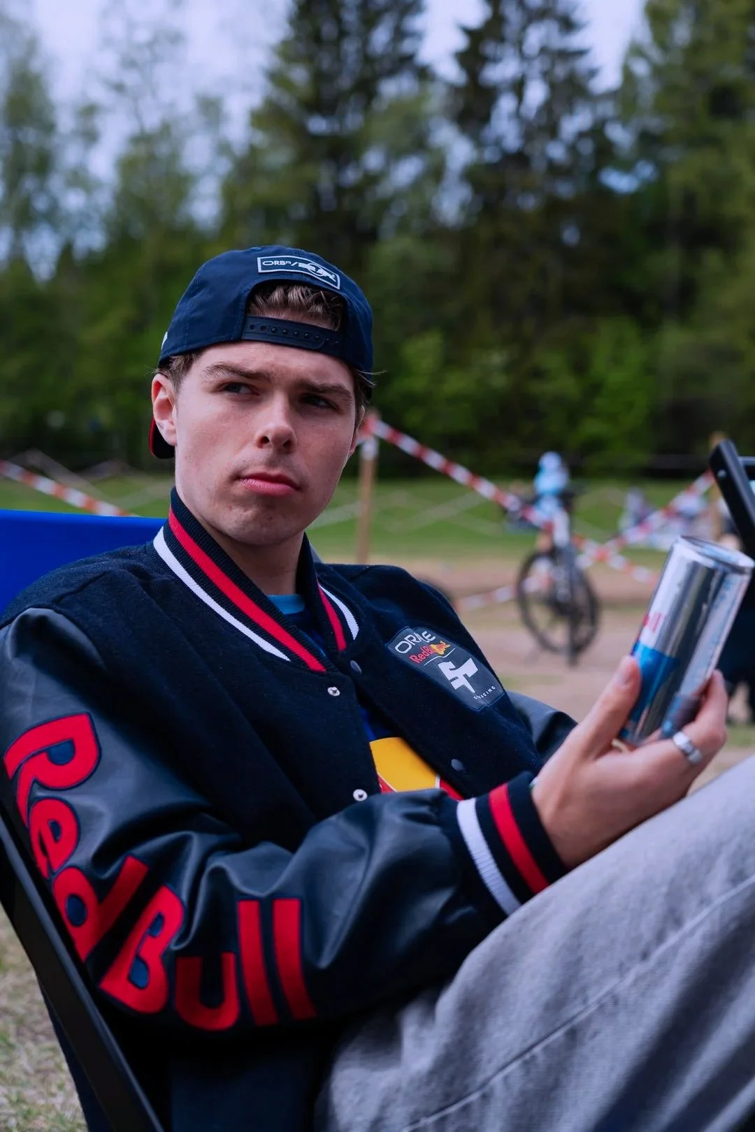 A young man sitting outdoors in a wooded area, wearing a Red Bull Racing jacket and a backwards cap, looking at his phone with a serious expression. In the background, there is a bicycle and some people, with red and white striped barrier tape.