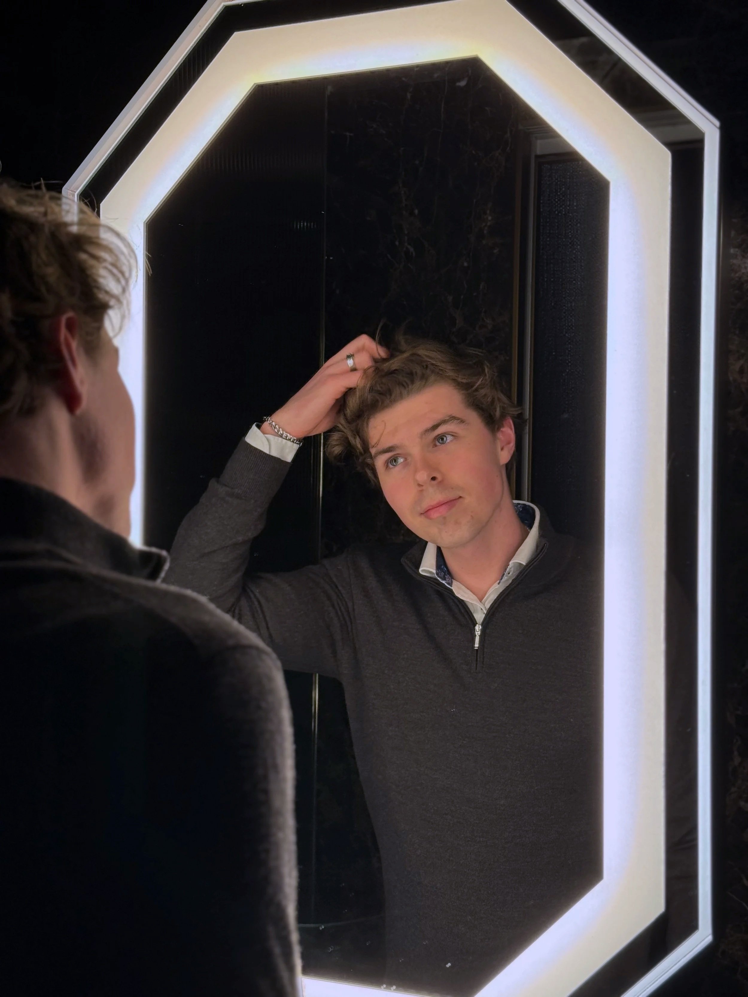A young man with curly hair looks at his reflection in a mirror with LED lighting, while touching his hair with one hand.