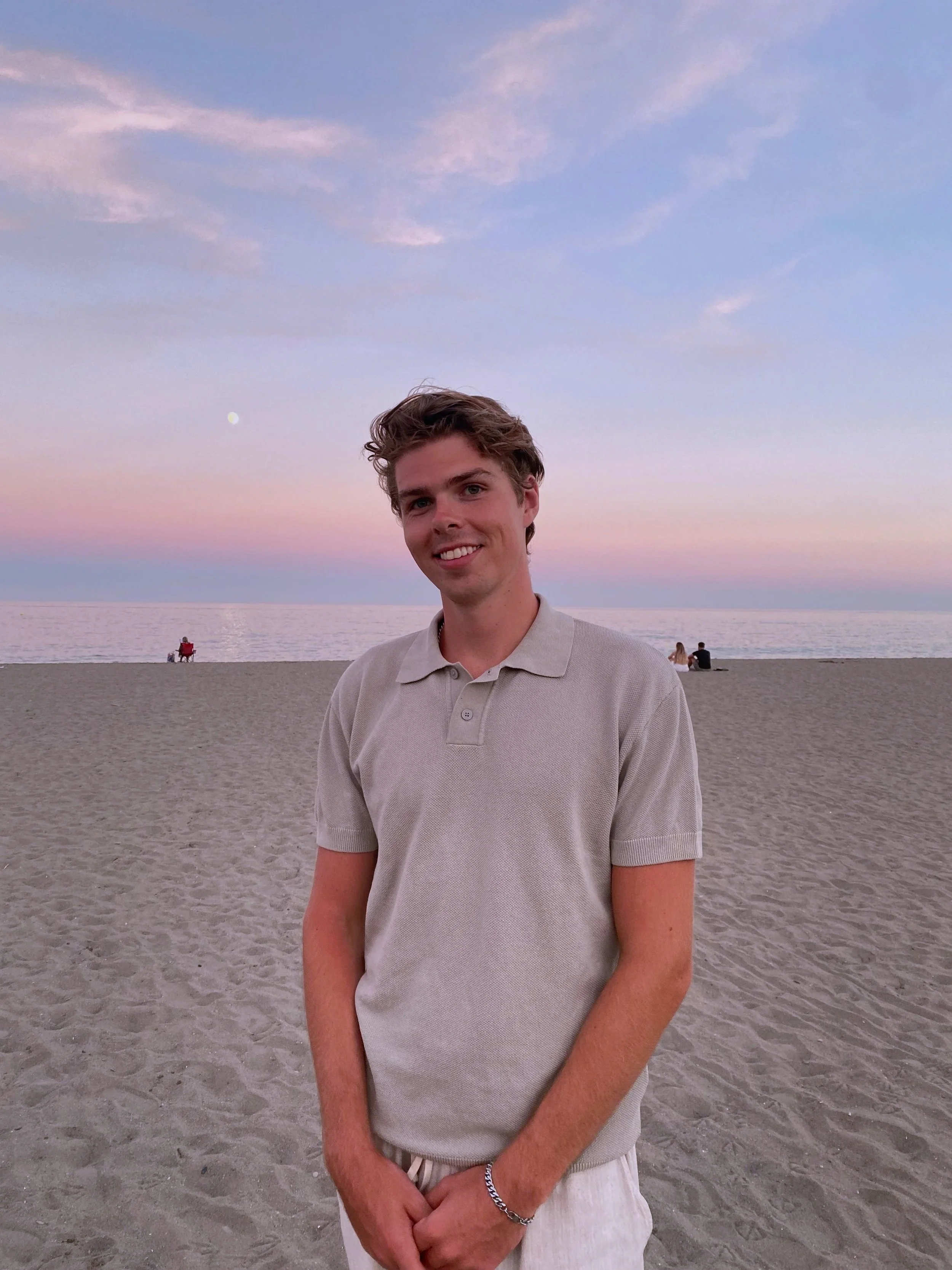 Young man with light brown hair smiling on a sandy beach during sunset with the ocean and a pink and blue sky in the background.