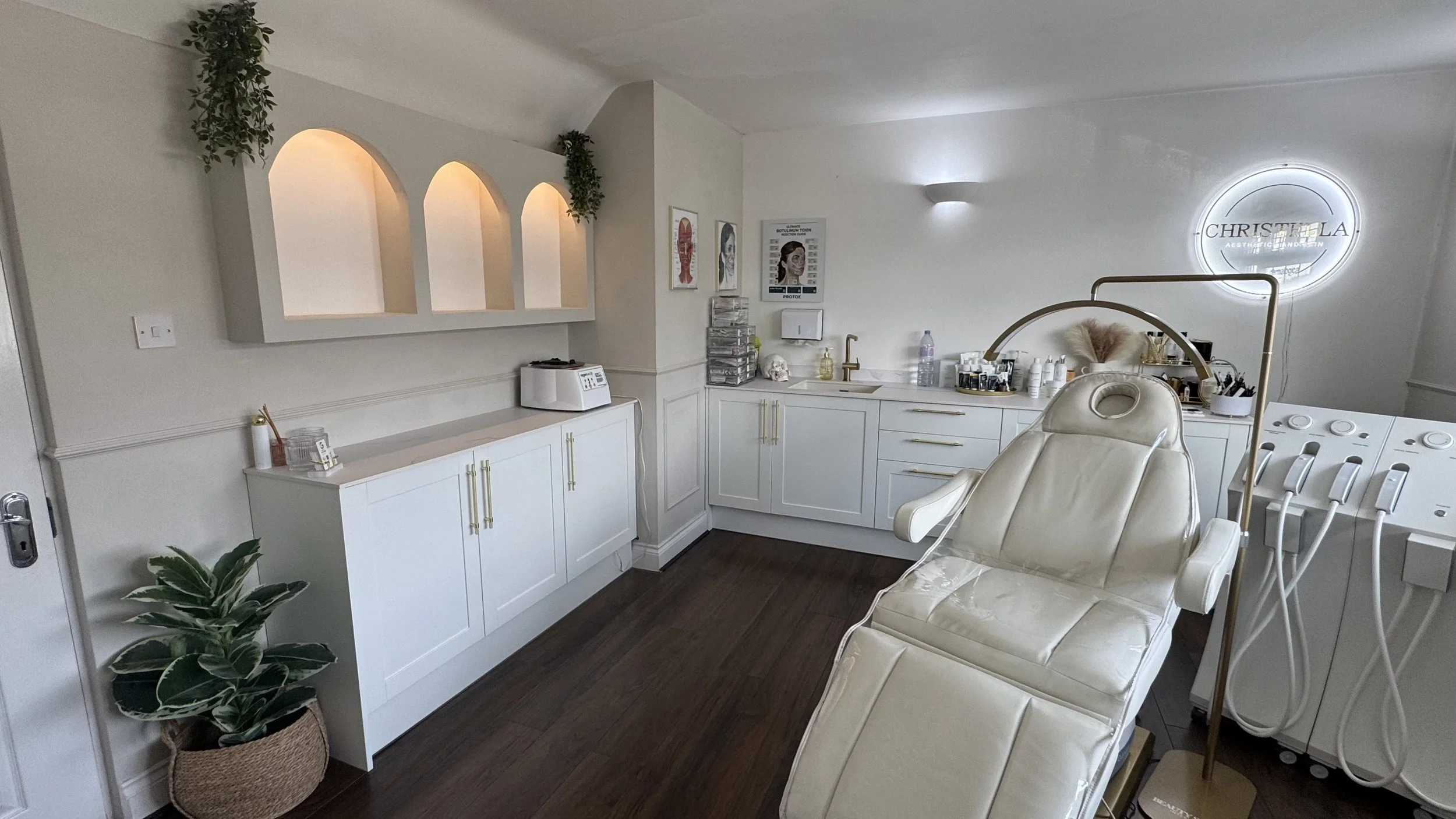 A medical or beauty clinic room with white cabinetry, a treatment chair, and medical or aesthetic supplies, including skincare products and medical posters on the wall.