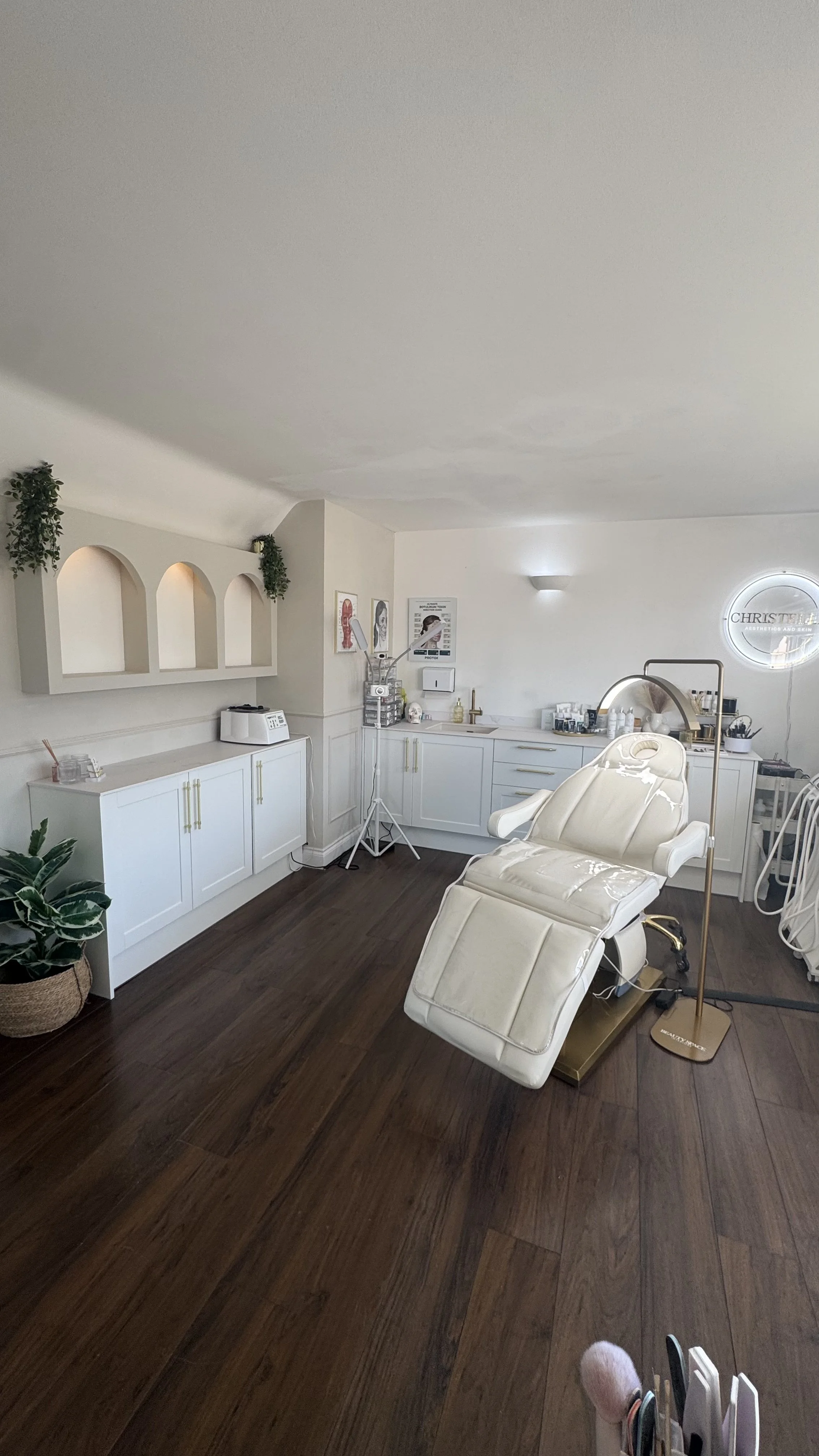 A bright and clean medical or skincare treatment room with white cabinetry, a reclining treatment chair, and equipment like a lamp and skincare products. There are decorative wall arches with greenery and framed pictures.