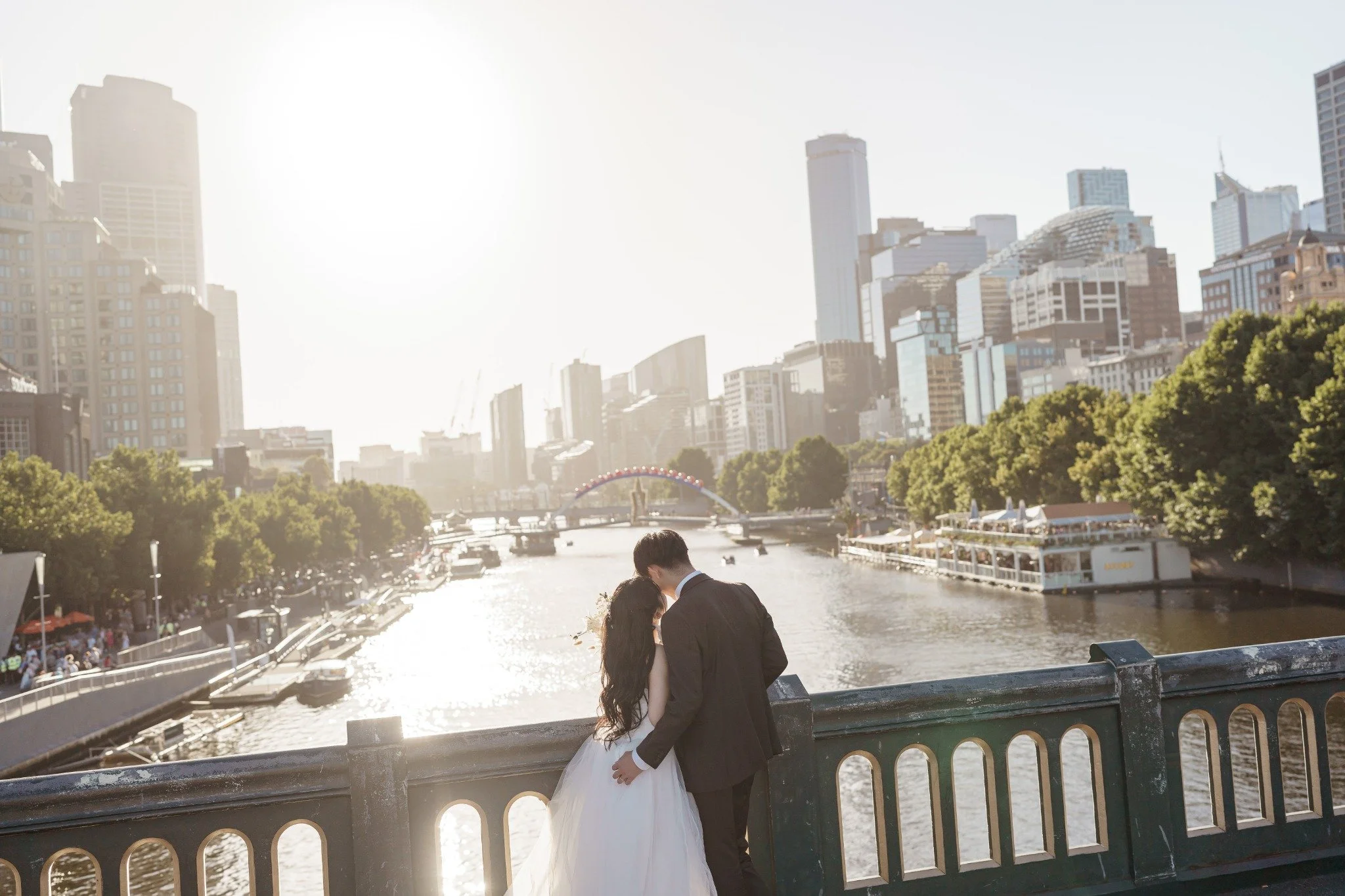 A bride and groom standing close together on a bridge, overlooking a river with boats, city skyline in the background, and the sun shining brightly.