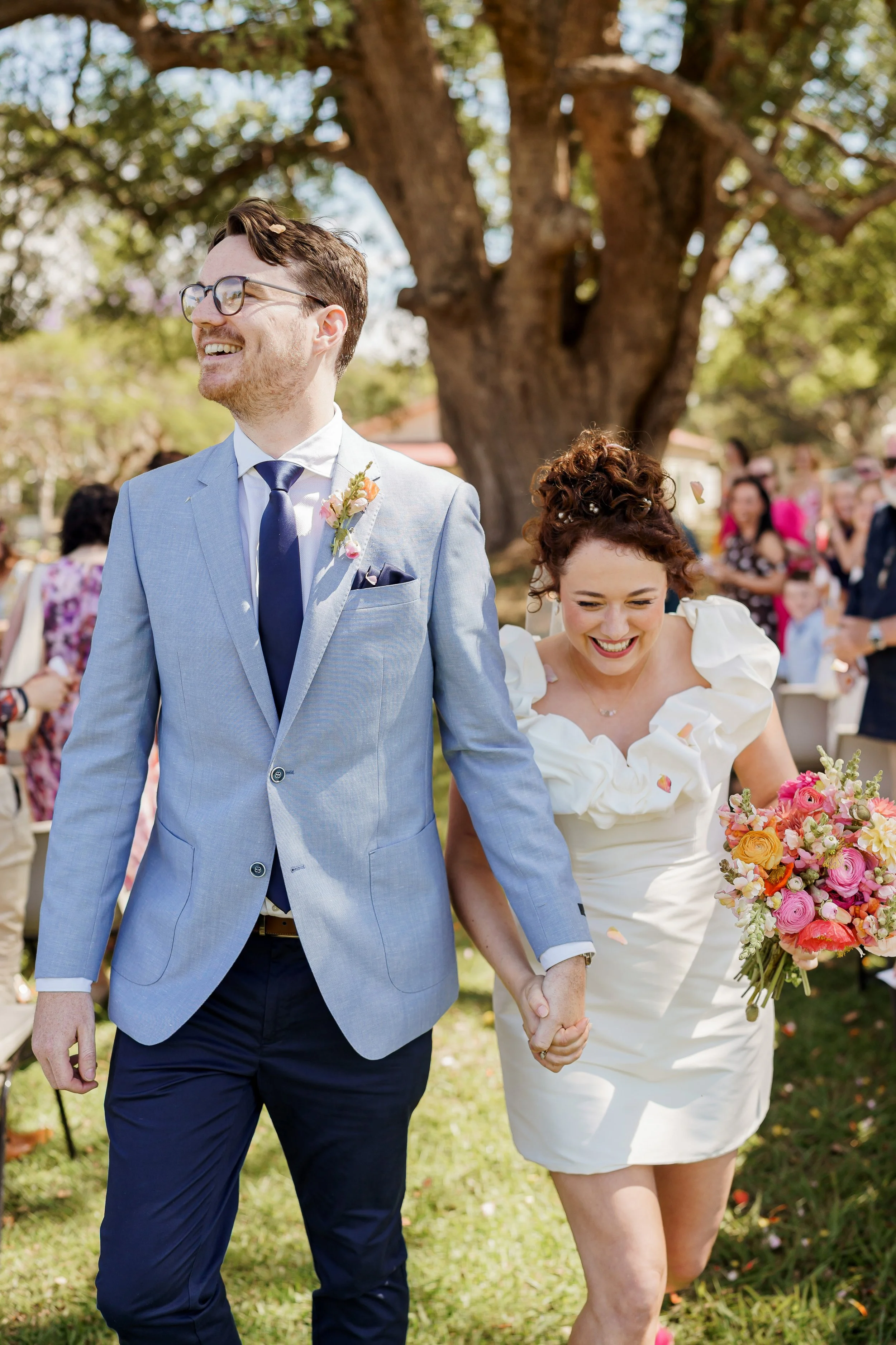 A smiling couple, dressed in wedding attire, walks hand-in-hand outdoors during a sunny day. The man is wearing a light blue suit with a dark tie and glasses, and the woman is in a white dress holding a bouquet of colorful flowers. There are people i