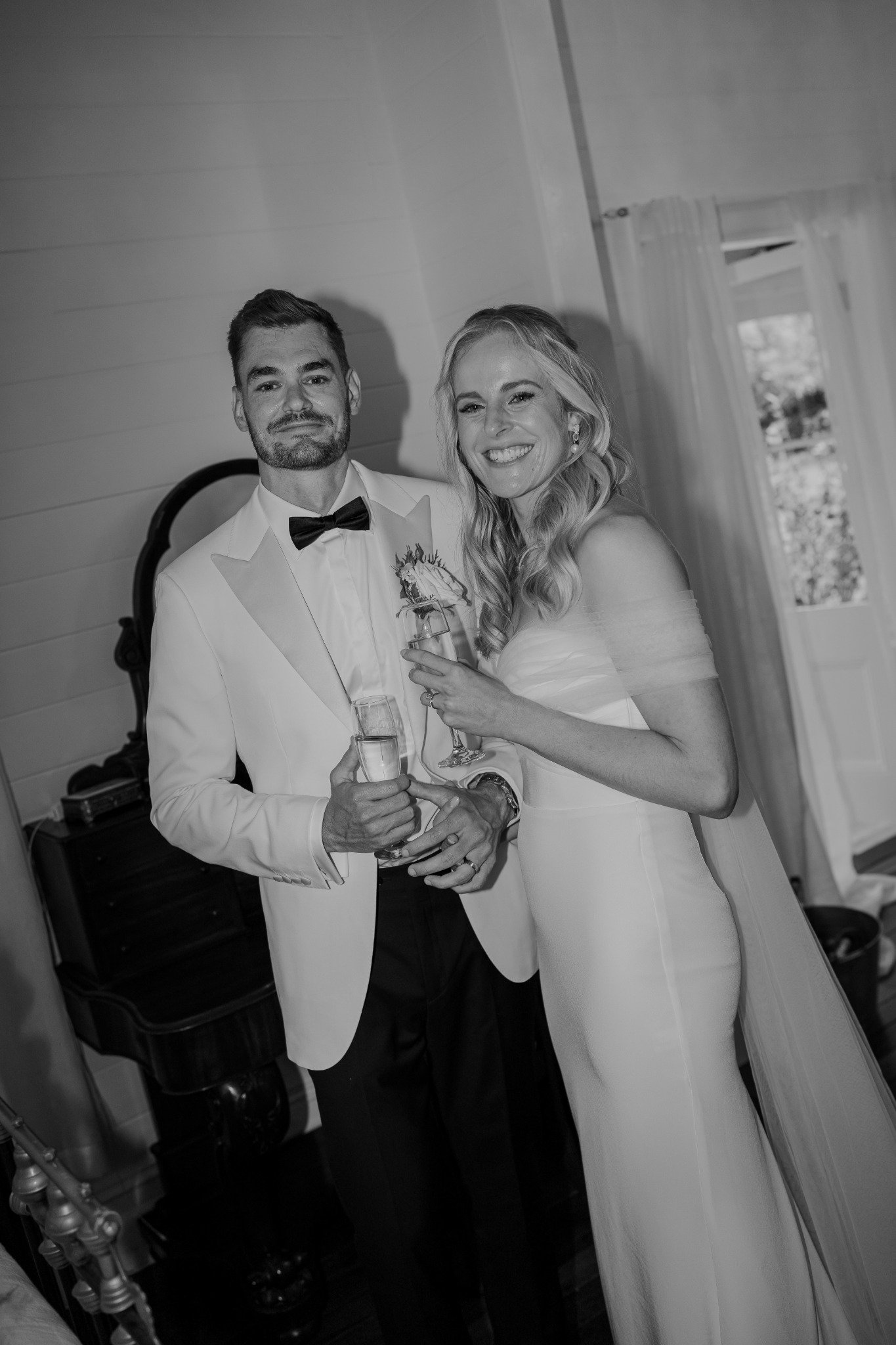A black and white photo of a couple in wedding attire posing together, each holding a glass of champagne, with the woman smiling and the man with a neutral expression inside a house with a window and curtain in the background.