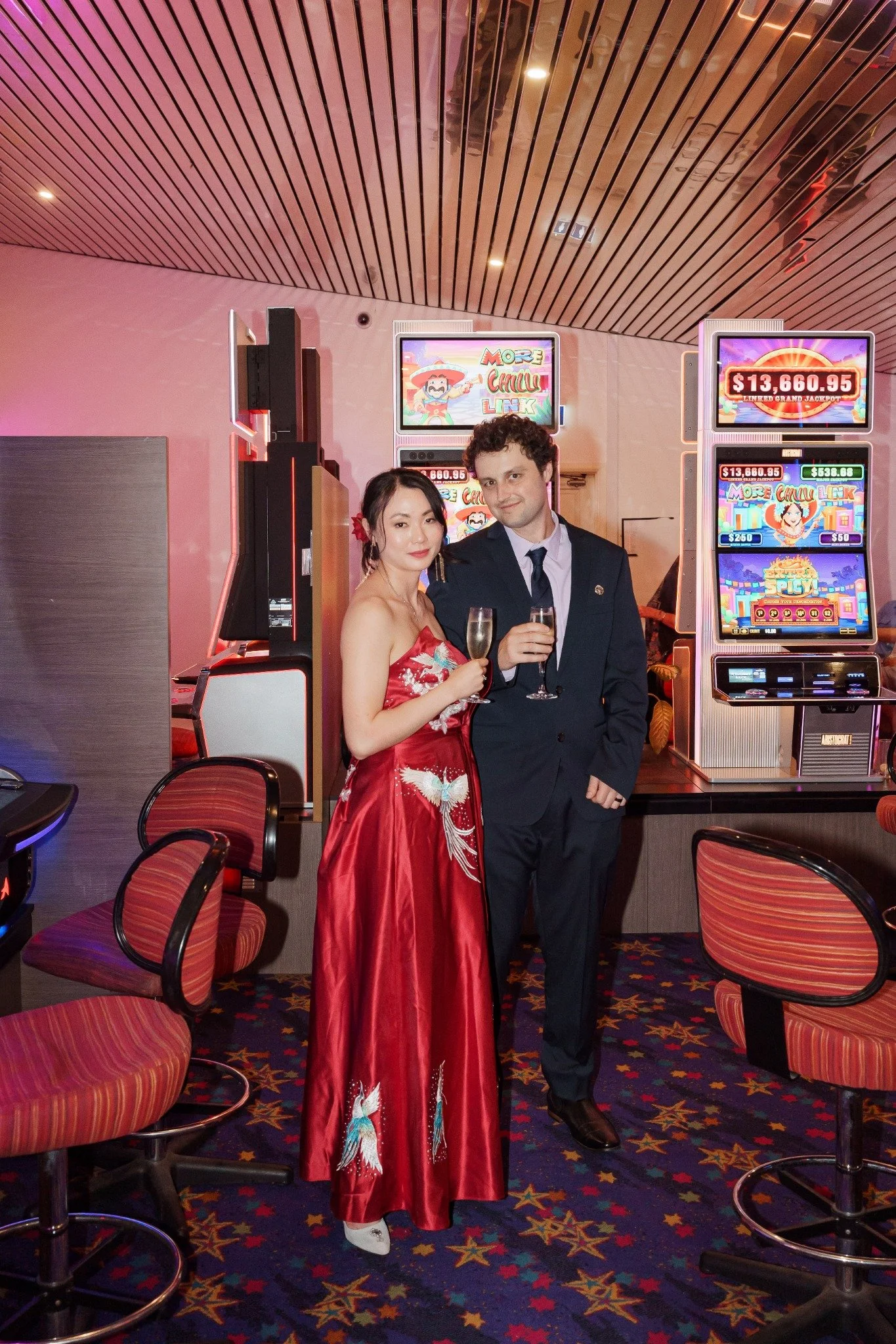 A woman in a red dress and a man in a suit holding glasses of champagne stand in a casino, with slot machines and a colorful carpet in the background.
