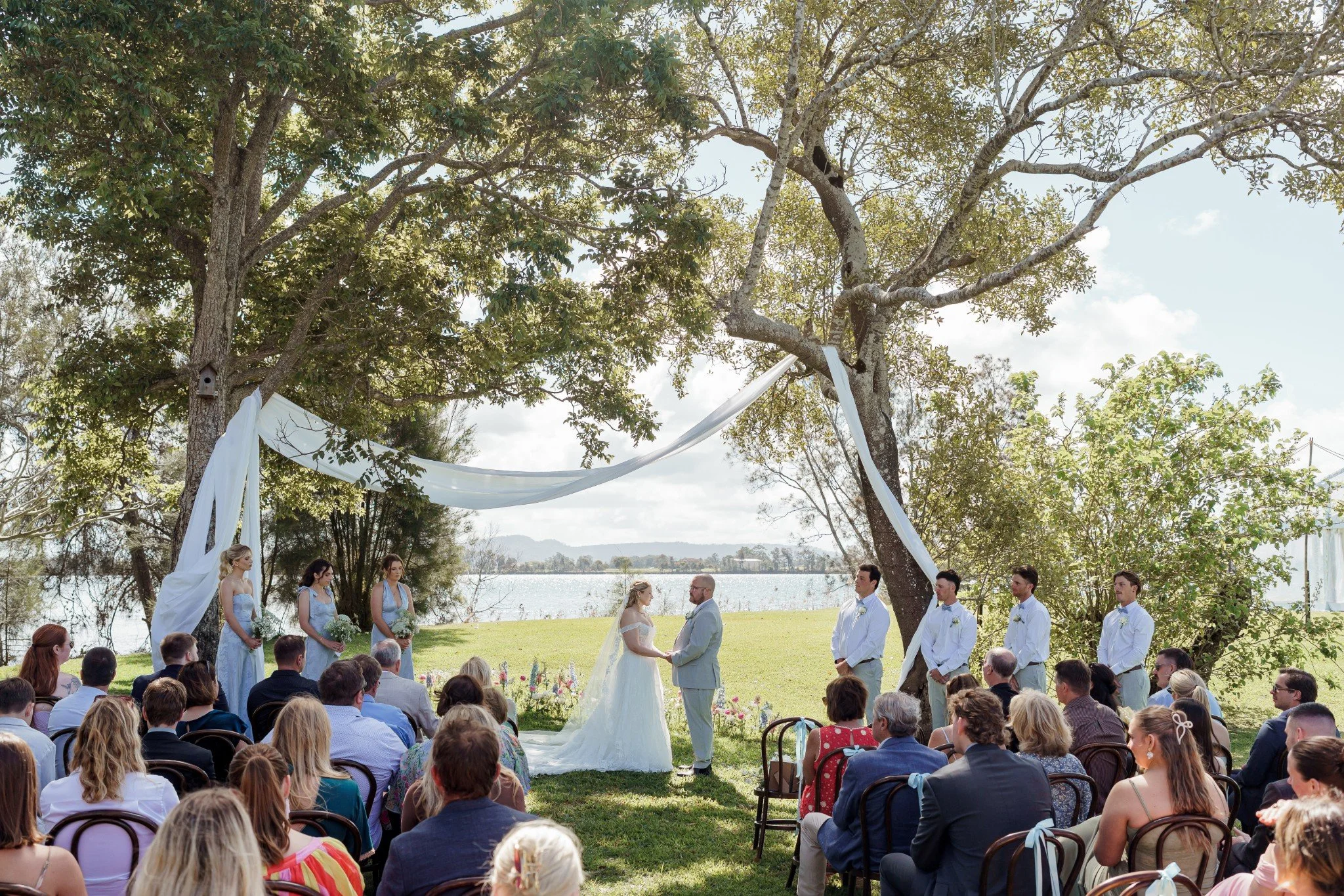 Outdoor wedding ceremony at Wynyabbie House in Yamba, with the bride and groom exchanging vows beneath trees overlooking the water.