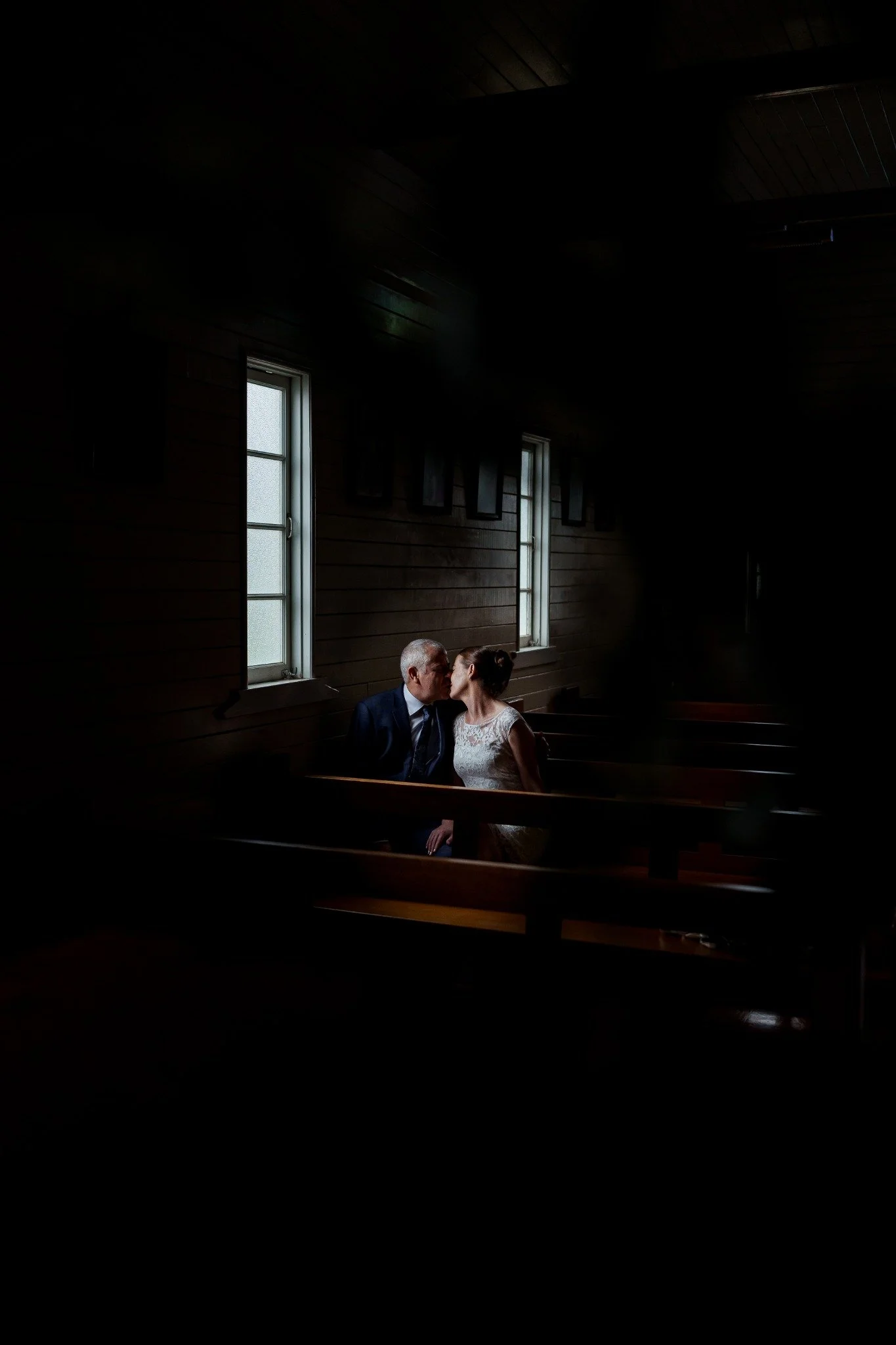 An elderly man and a young woman share a kiss while seated in a church pew near two tall, narrow windows with frosted glass, in a dimly lit church with dark wooden walls.