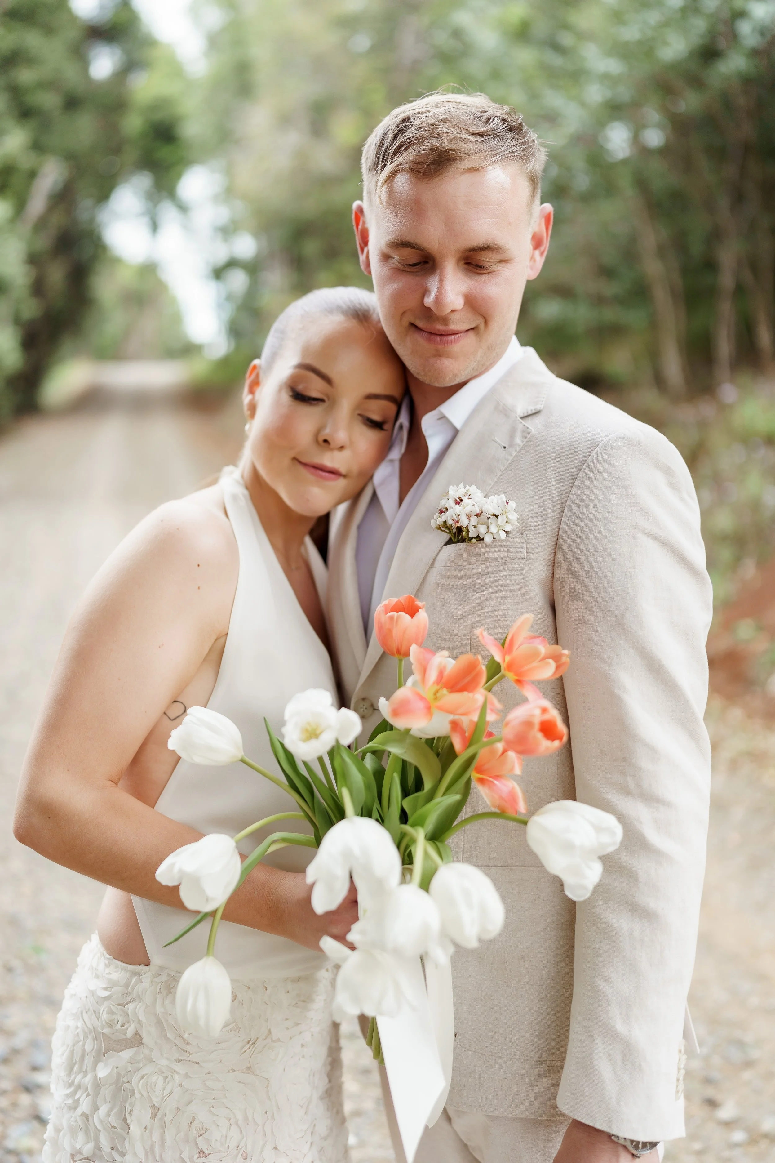 A bride and groom in wedding attire embracing outdoors, holding a bouquet of pink, white, and orange tulips, with a green wooded background.