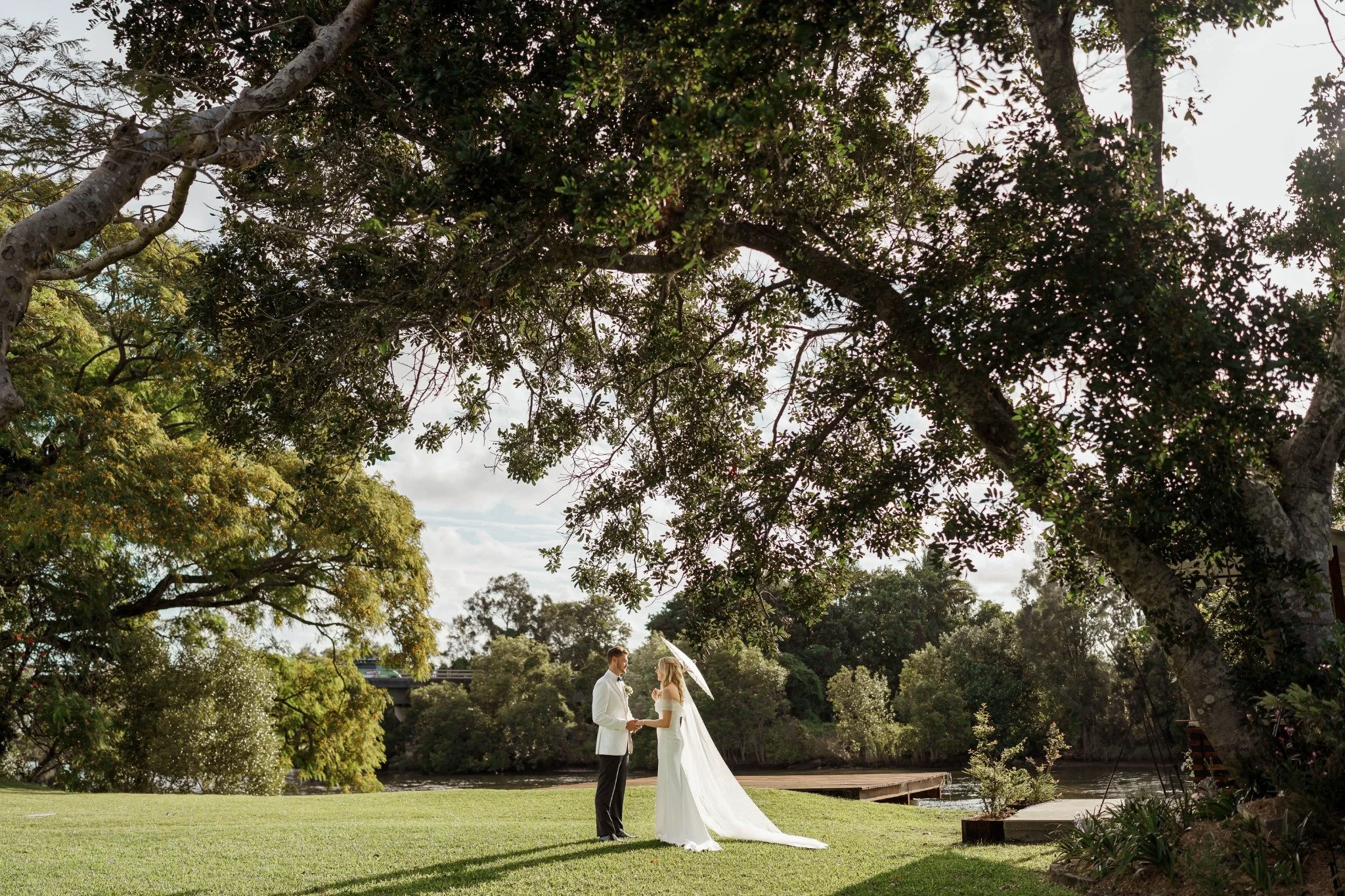 A bride and groom standing under a large tree, holding hands and facing each other, with a river and trees in the background. The bride is holding a white umbrella and wearing a white wedding dress and veil.
