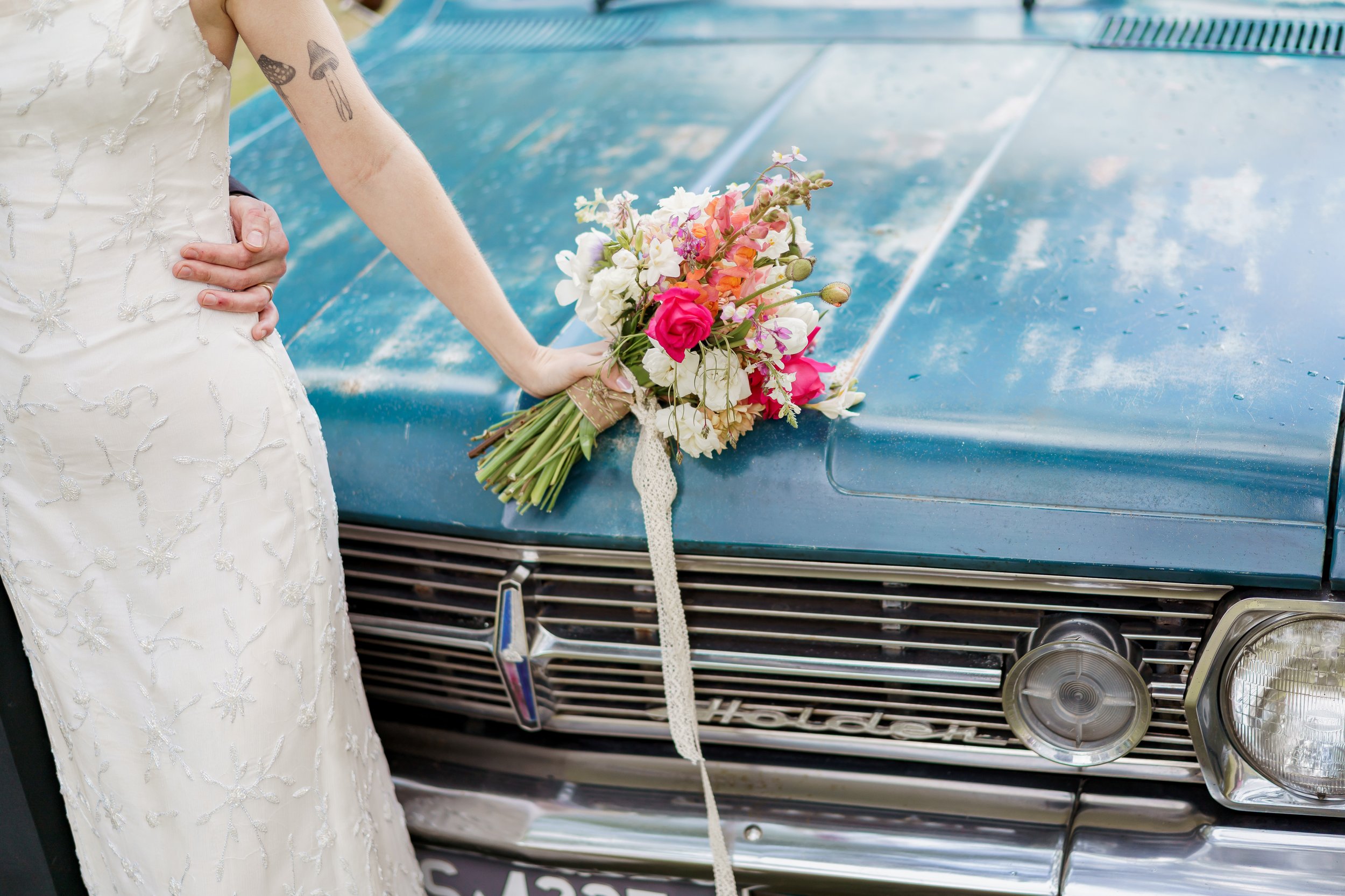 A bride in a white dress with floral embroidery is holding a bouquet of pink, white, and purple flowers on the hood of a vintage blue car decorated with a floral ribbon.