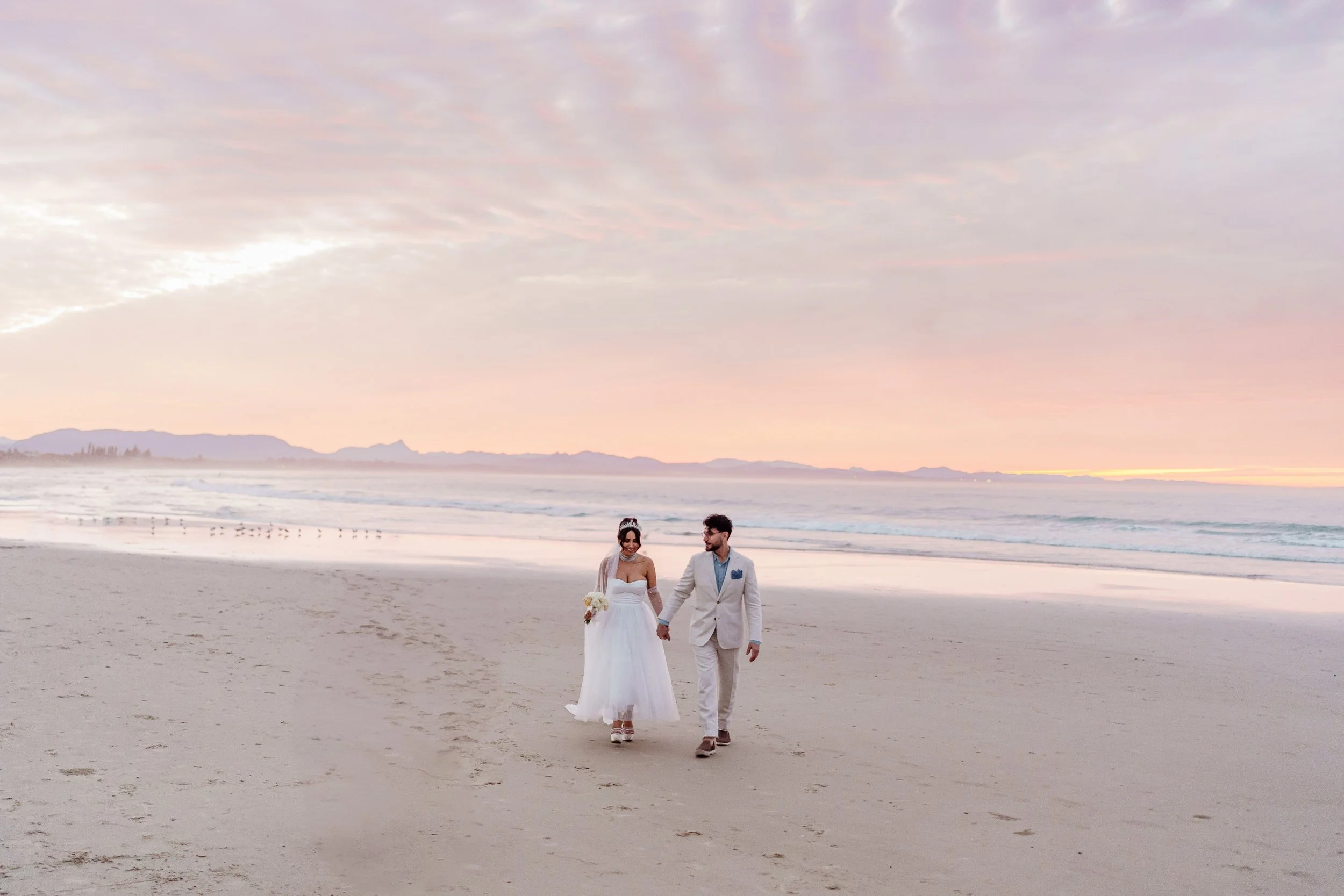 A bride and groom holding hands and walking on the beach at sunset, with the ocean and distant mountains in the background.