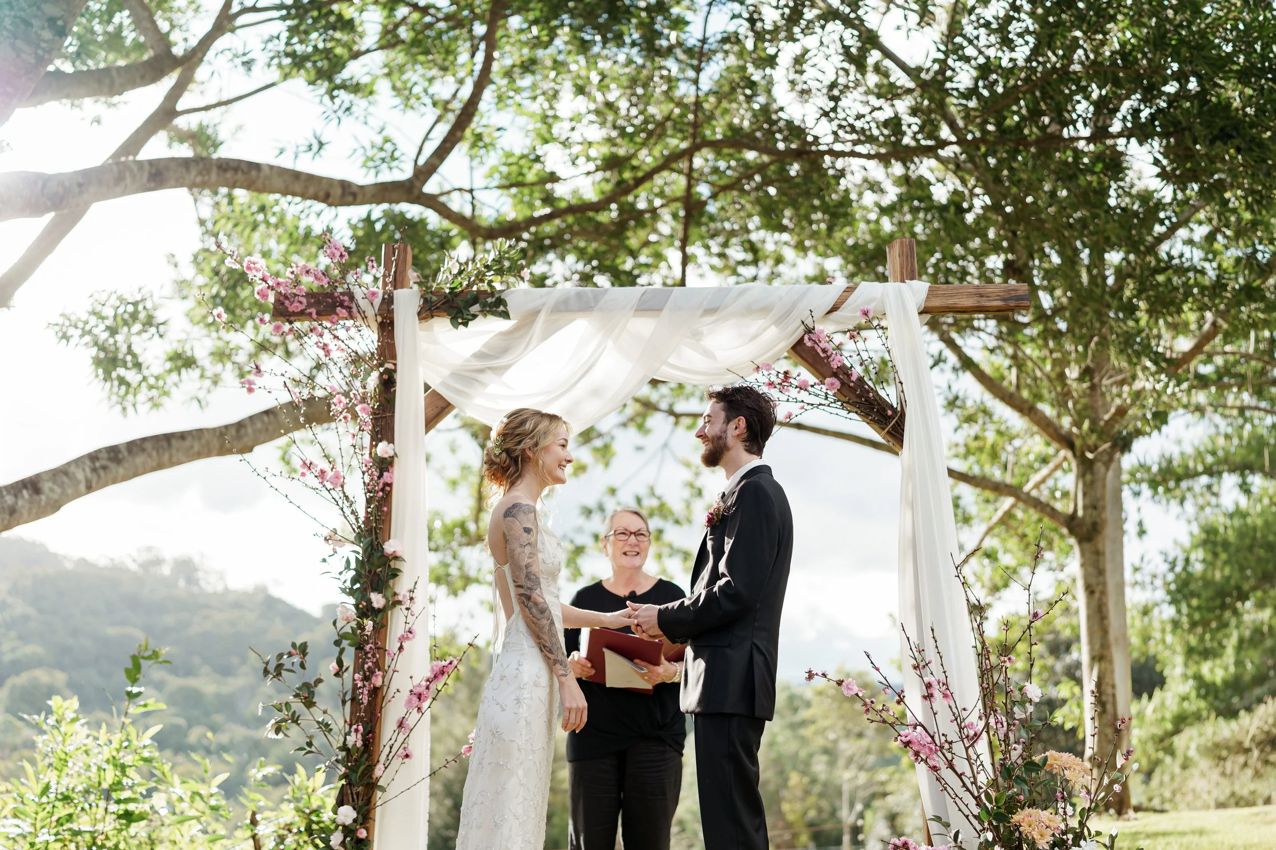 A couple getting married outdoors under a floral wedding arch, holding hands and smiling at each other as an officiant looks on.