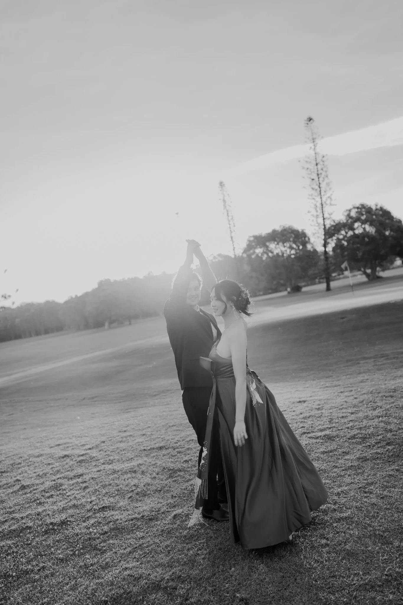 A couple dancing on a grassy field with trees in the background, backlit by the setting or rising sun.