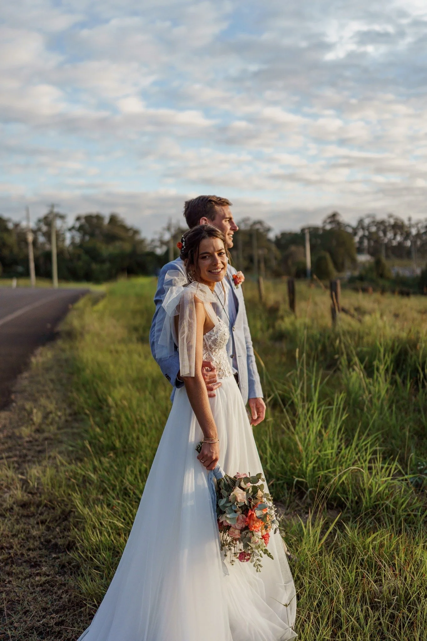 A bride and groom standing outdoors on a grassy roadside, smiling and looking into the distance, with the bride holding a bouquet of flowers, during sunset or late afternoon.