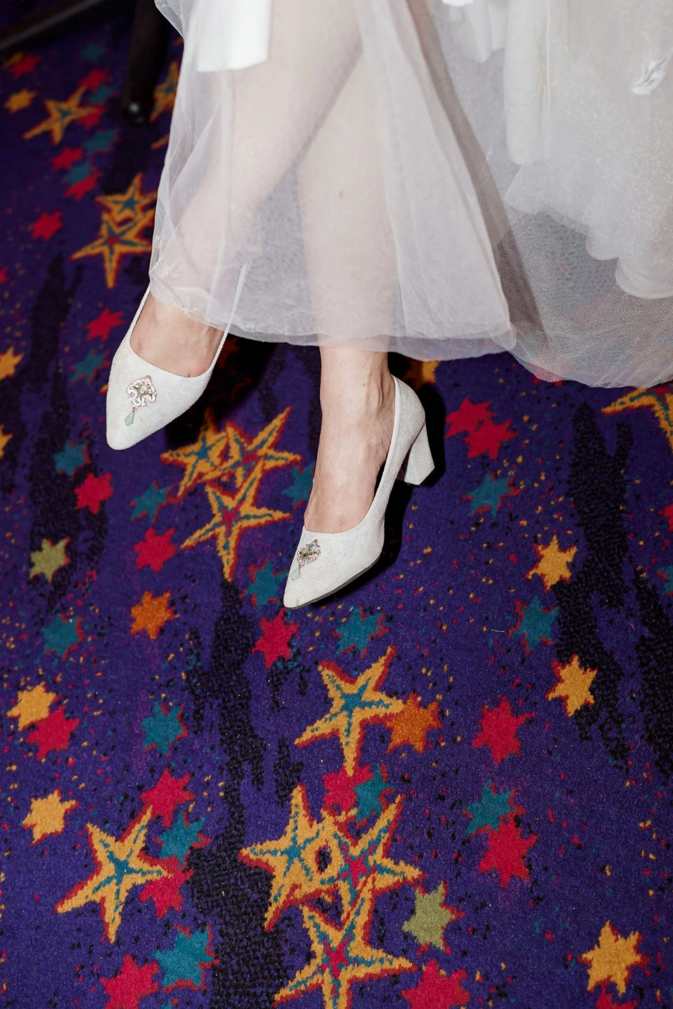 Close-up of a woman's feet in white high heels with embellishments, standing on a colorful patterned carpet beneath a semi-transparent white dress.