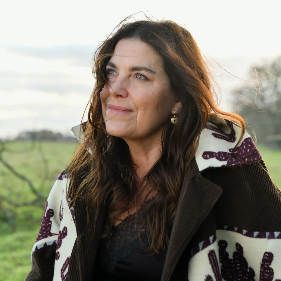 A woman with long brown hair and blue eyes looks to the side outdoors, wearing earrings and a patterned coat, with green fields and cloudy sky in the background.