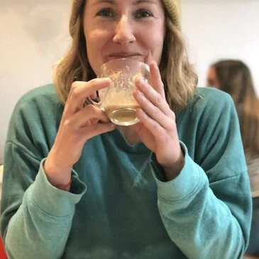 Person sitting, smiling while holding a coffee cup, wearing a beanie and teal sweatshirt.