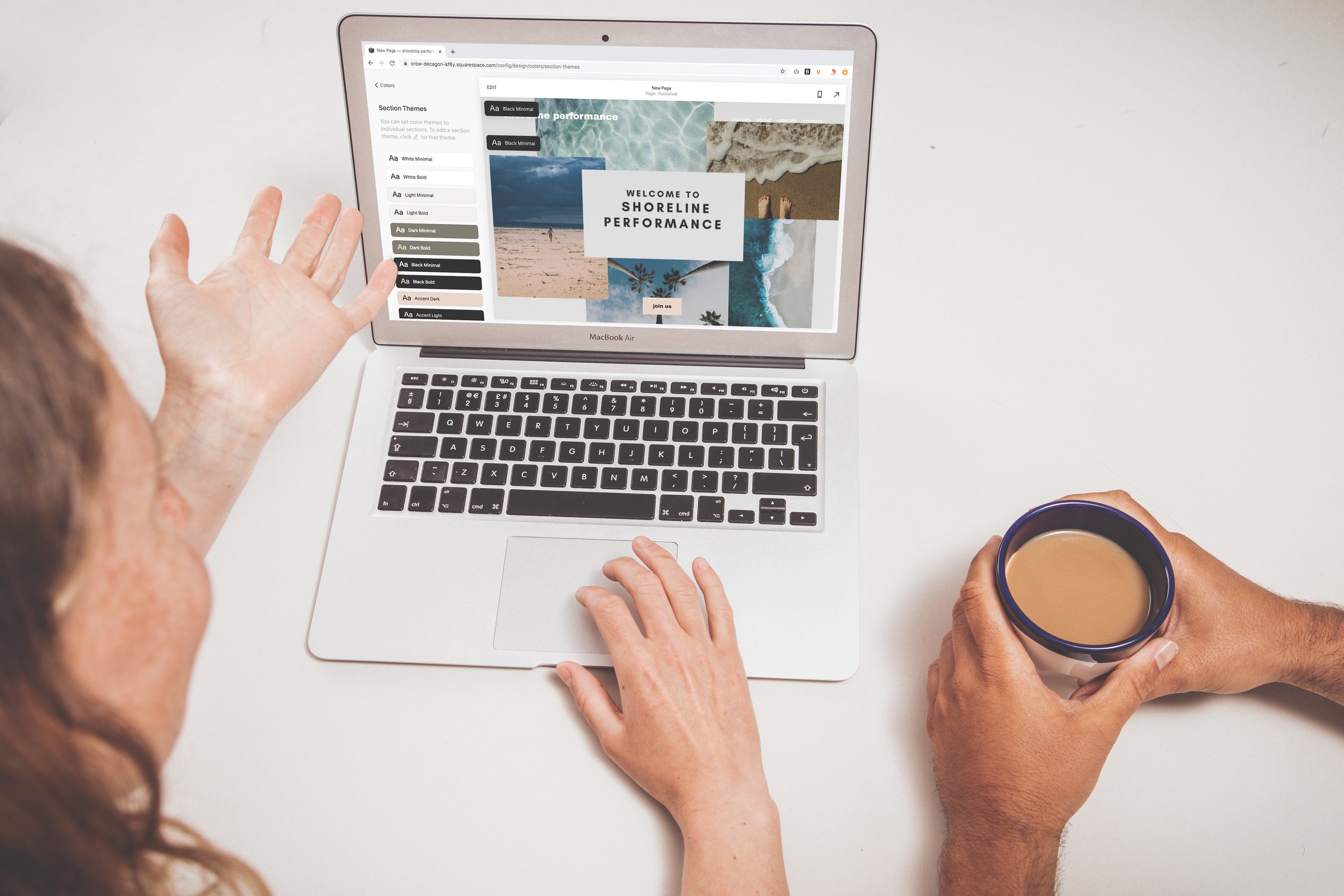 Two people reviewing a website design on a MacBook Air, one gesturing and the other holding a coffee mug.