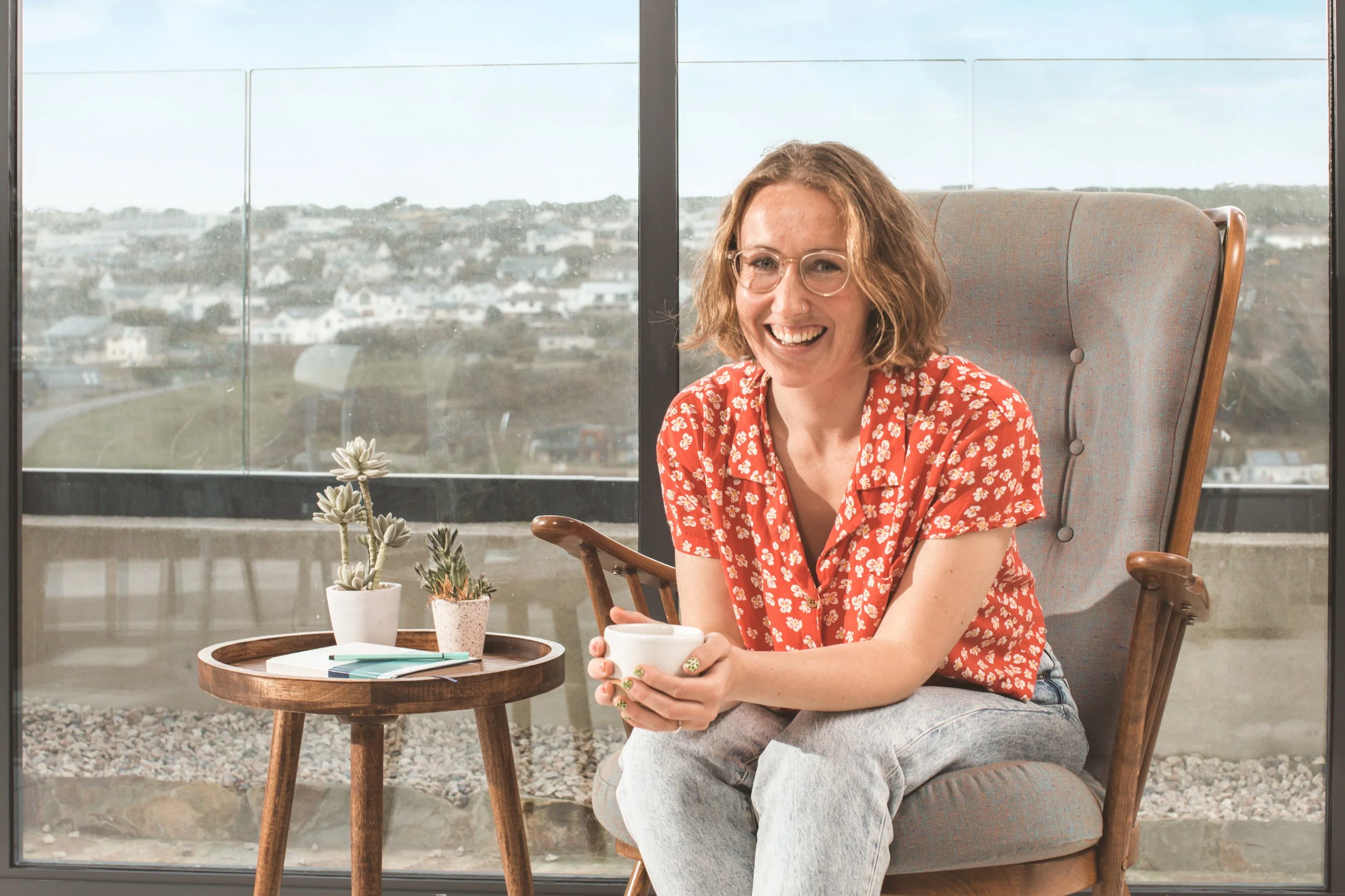Smiling woman in glasses and red floral shirt sitting with a cup in hand, next to a small table with succulents, in a cozy, sunlit room with city view.