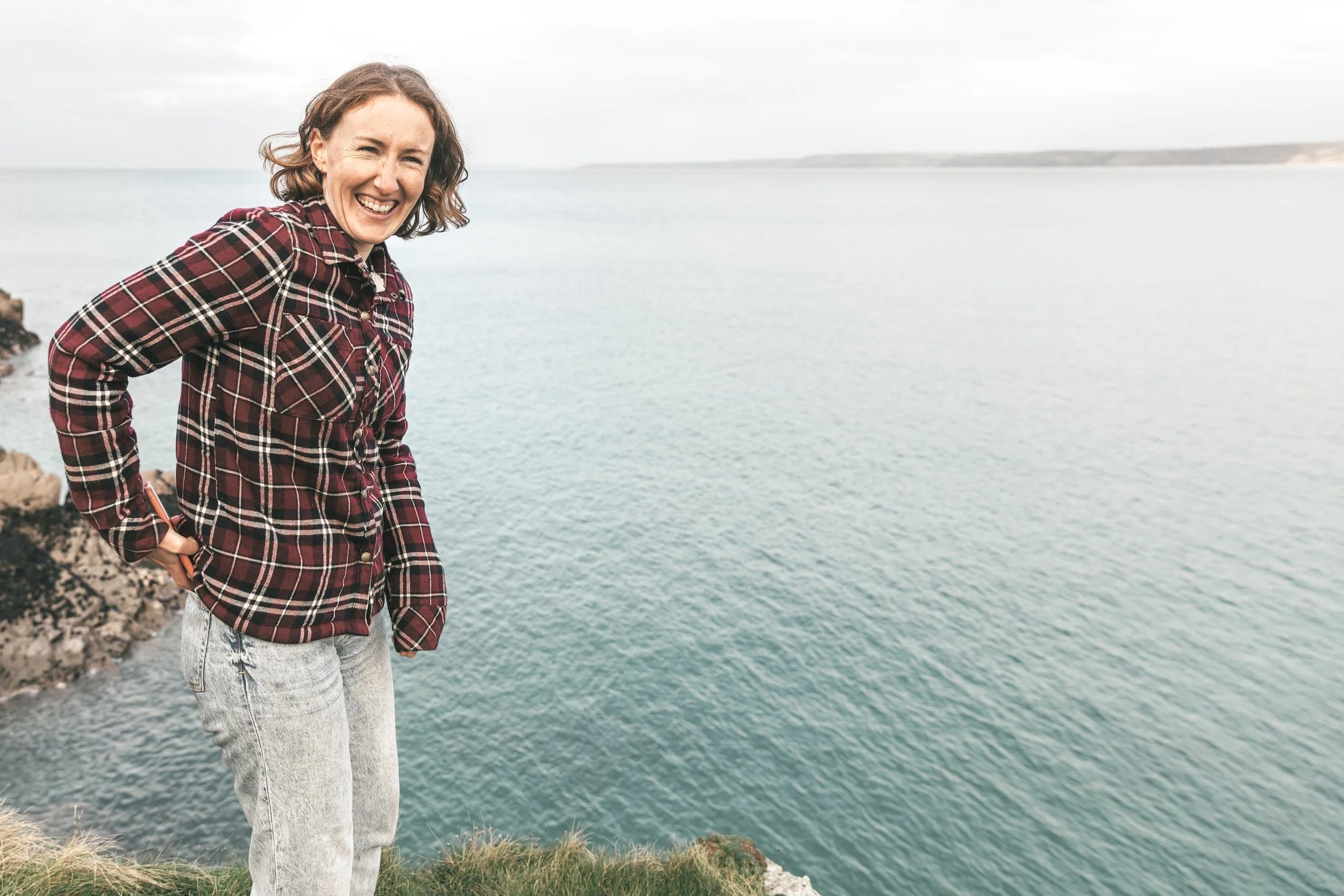 Person smiling, wearing a plaid shirt and jeans, standing near a cliff with the ocean in the background.