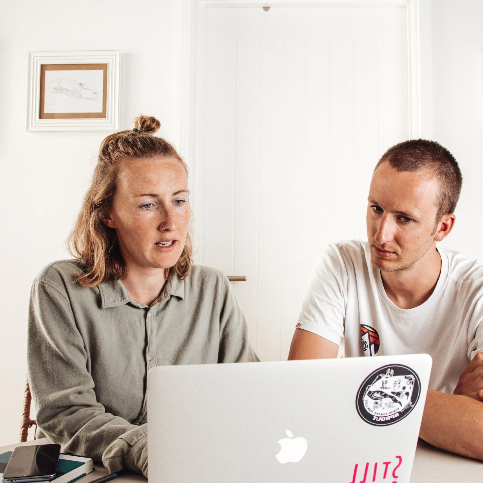 Two people sitting at a table, working on a laptop together. A smartphone and notebooks are on the table. The background features a white door and a framed picture on the wall.