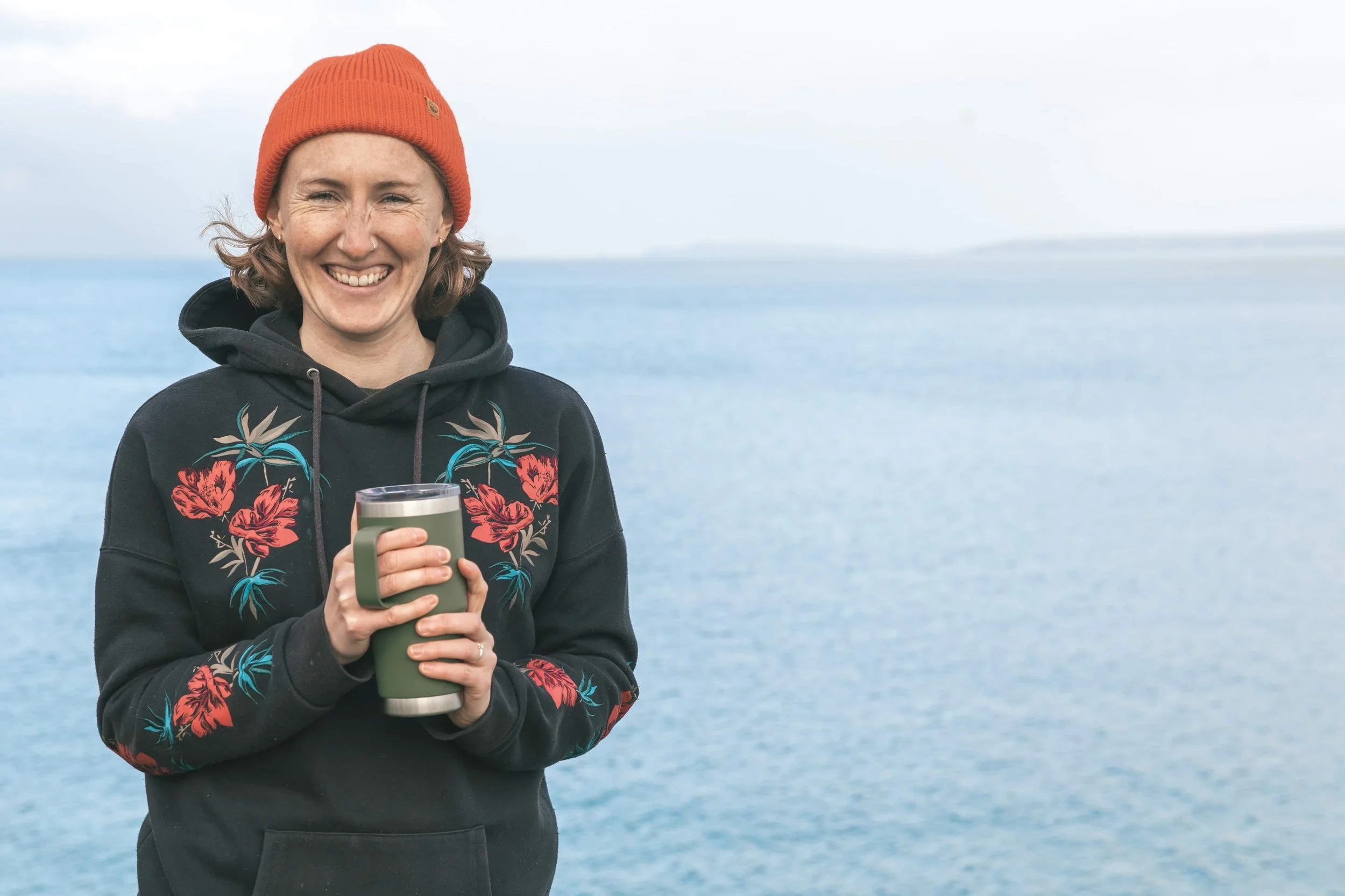 Person in a beanie and floral hoodie holding a mug by the ocean.