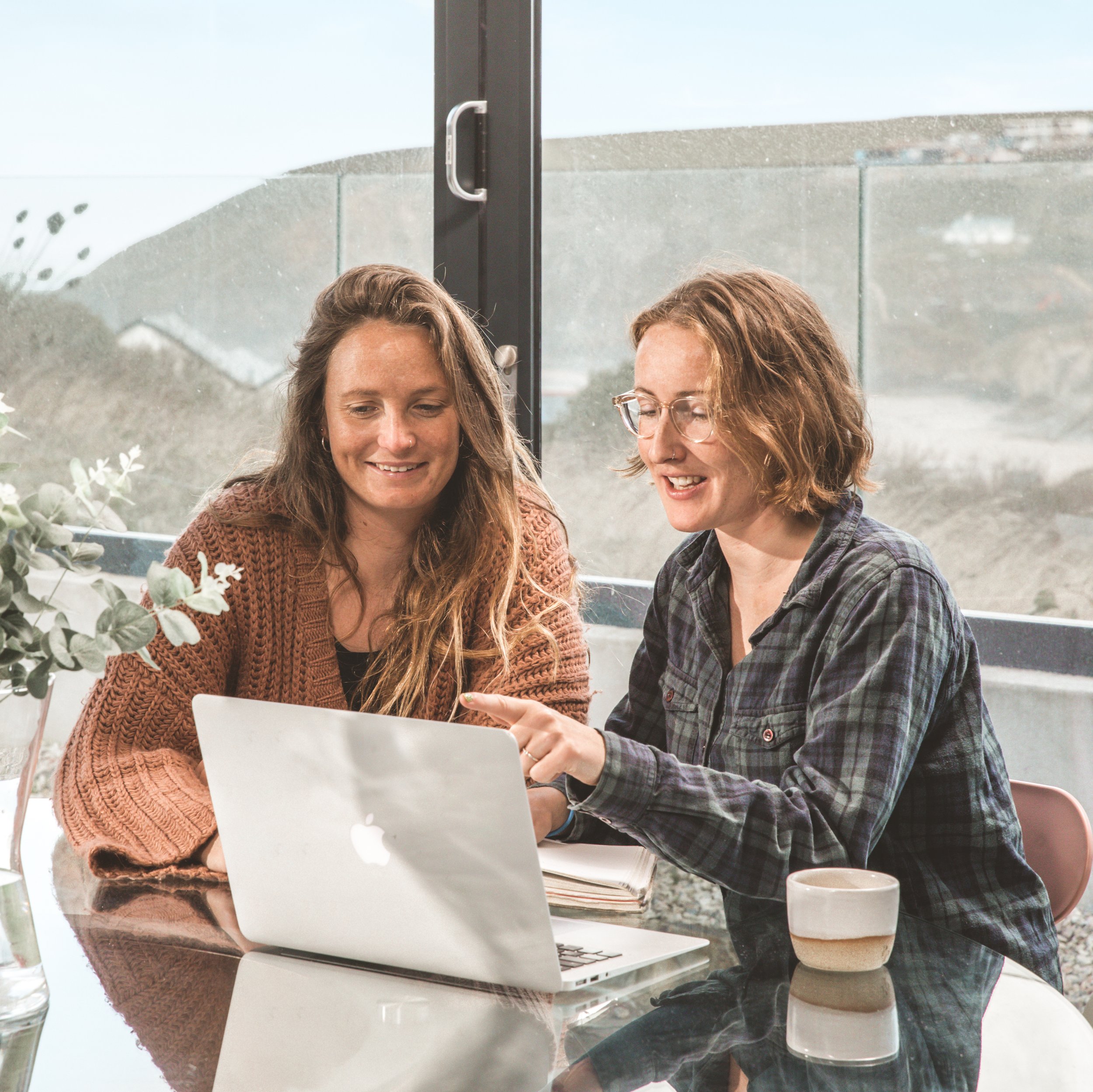 Two women sitting at a table with a laptop, looking at the screen and smiling, next to a window with an outdoor view.