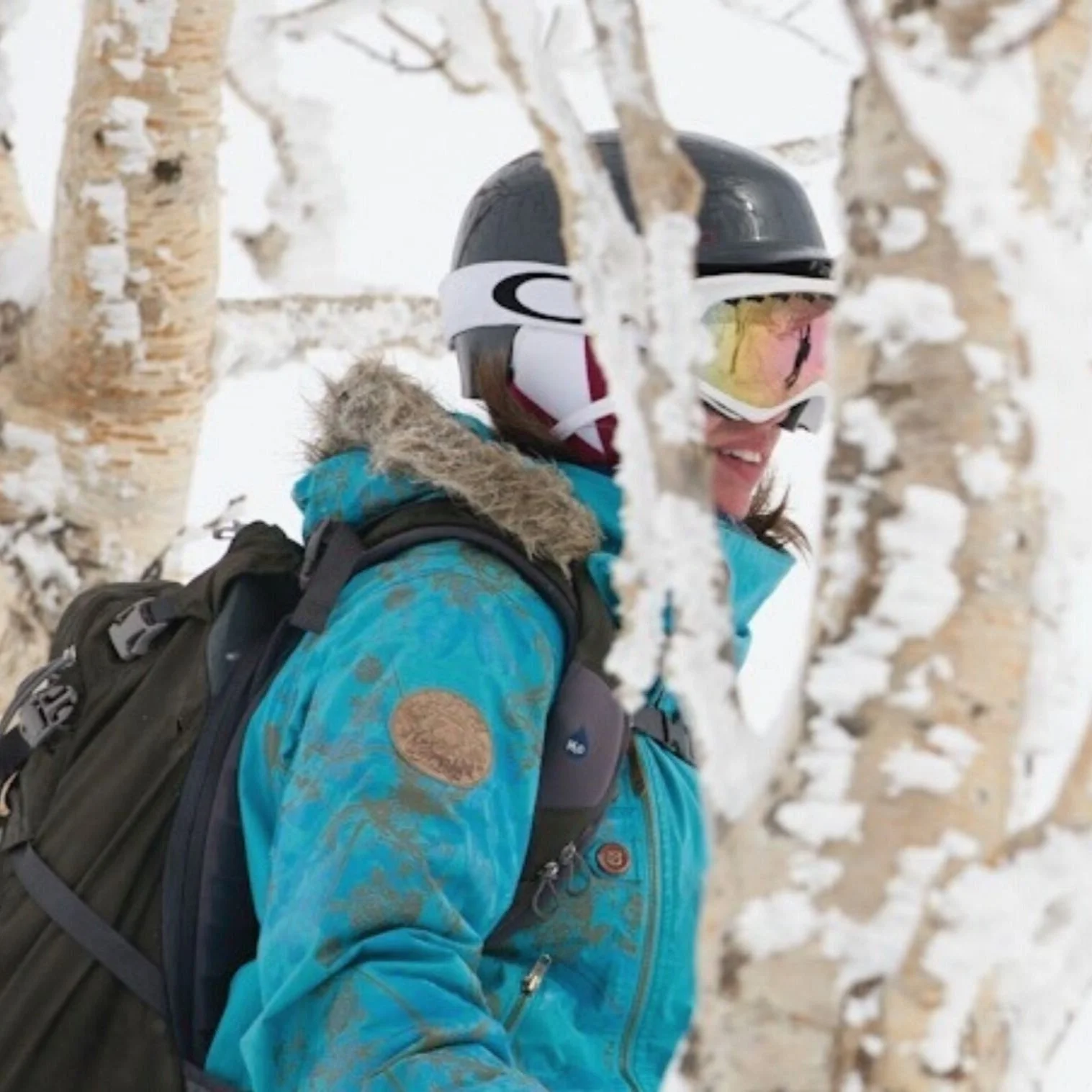 Person in blue jacket with fur hood, wearing ski helmet and goggles, carrying a backpack, standing among snow-covered trees.