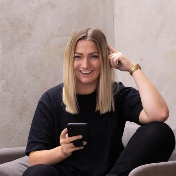Person sitting on a chair holding a smartphone, smiling, and wearing a black shirt and black pants.