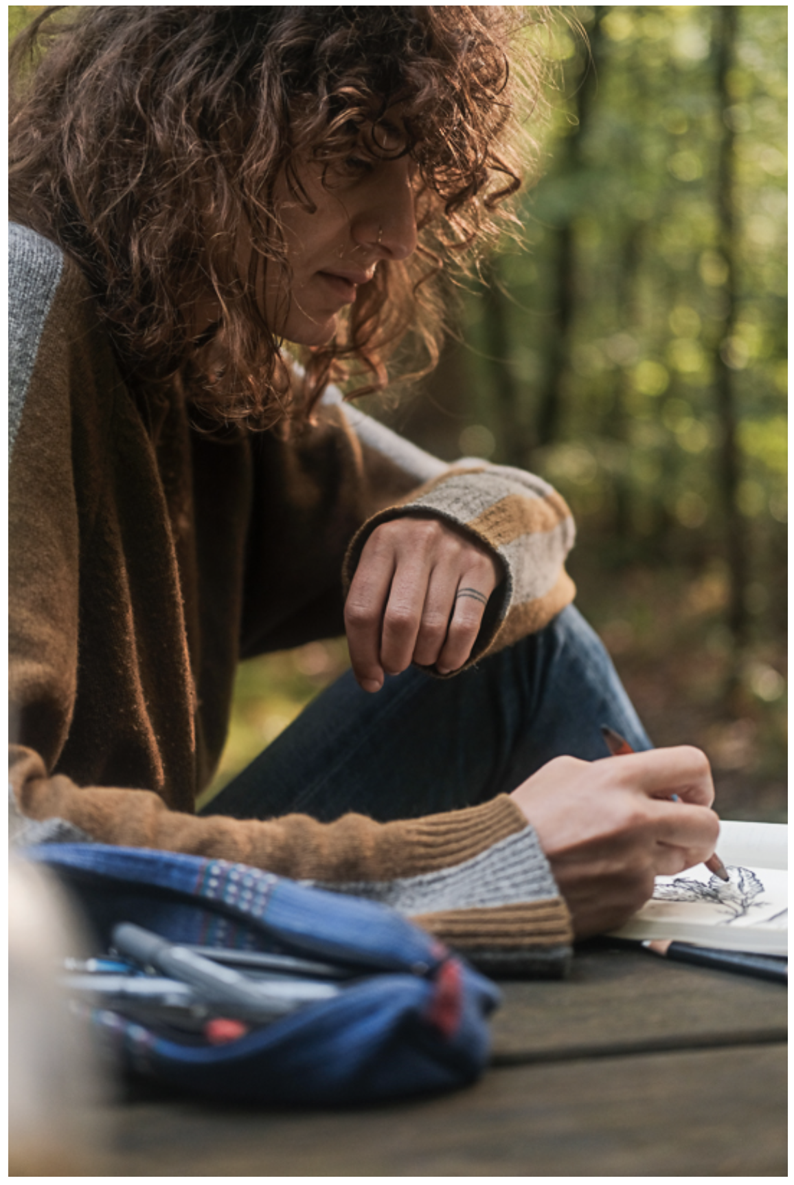 non-binary person sitting at a picnic bench in the forest looking down at their sketchbook while they draw in it with pencils
