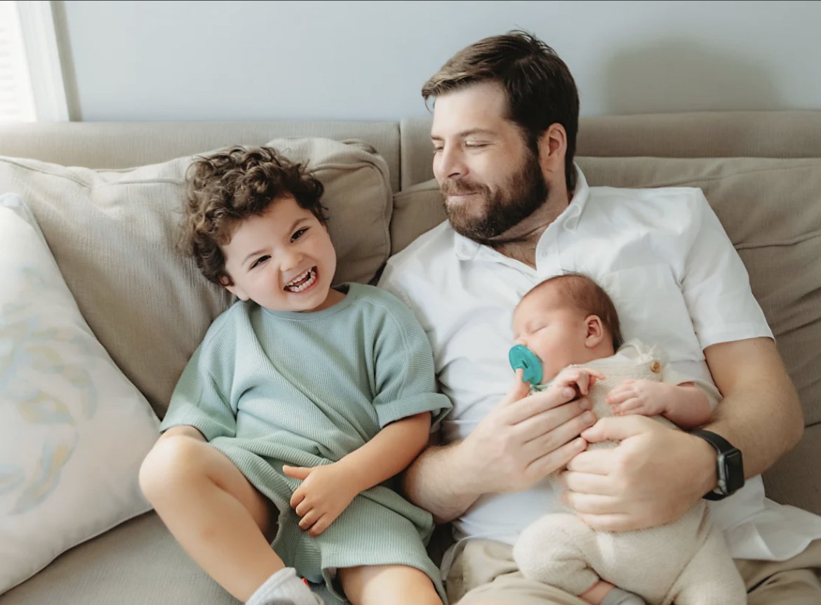 Jayme Marooney holding a sleeping baby with a pacifier, while a smiling, curly-haired older son sits beside them.