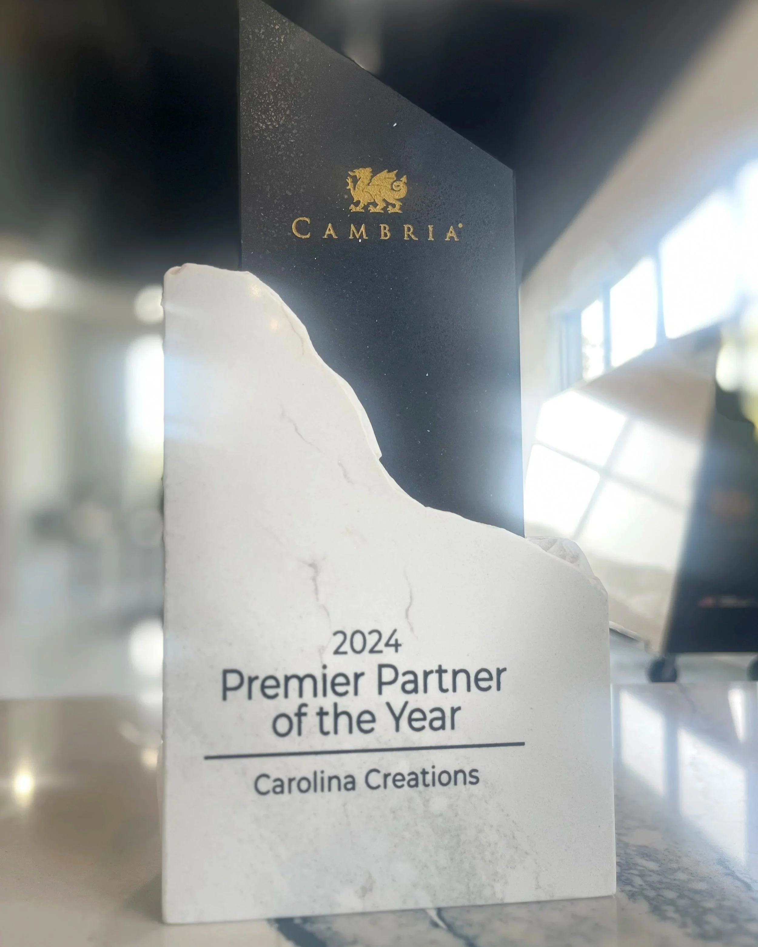 A white award plaque with the text '2024 Premier Partner of the Year, Carolina Creations,' and a black box with a gold logo and the word 'Cambria' behind it.