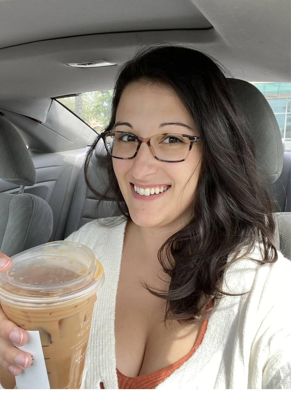 A woman with glasses and long dark hair smiling, holding an iced coffee, inside a car.