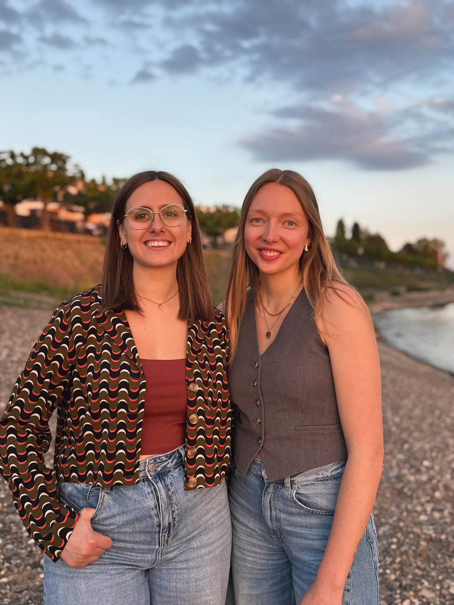 Zwei junge Frauen stehen am Strand bei Sonnenuntergang, lächeln und posieren für das Foto.