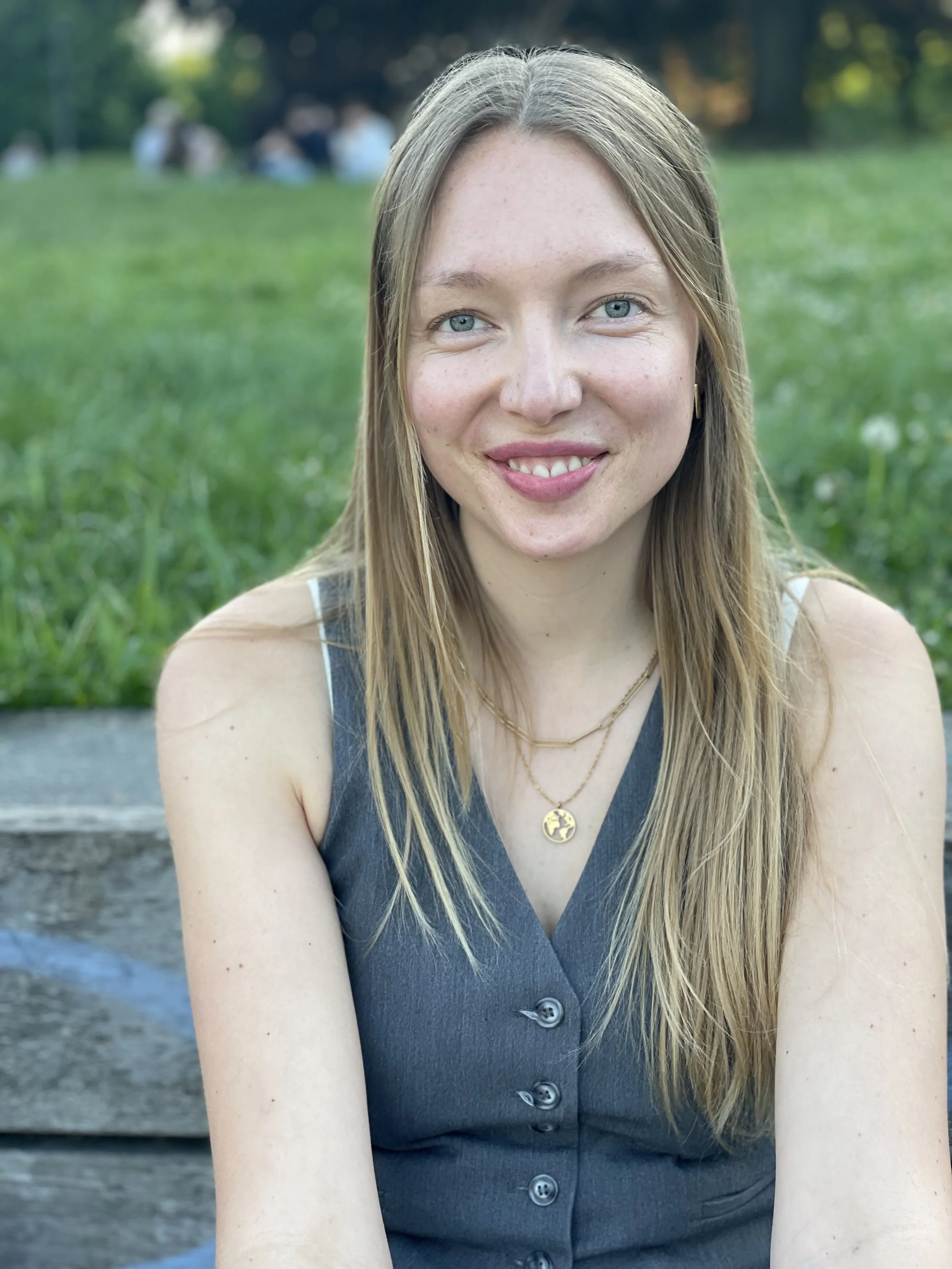 Ein junges Mädchen mit blonden Haaren, blauen Augen und einem Lächeln, sitzend im Freien auf einer grünen Wiese.