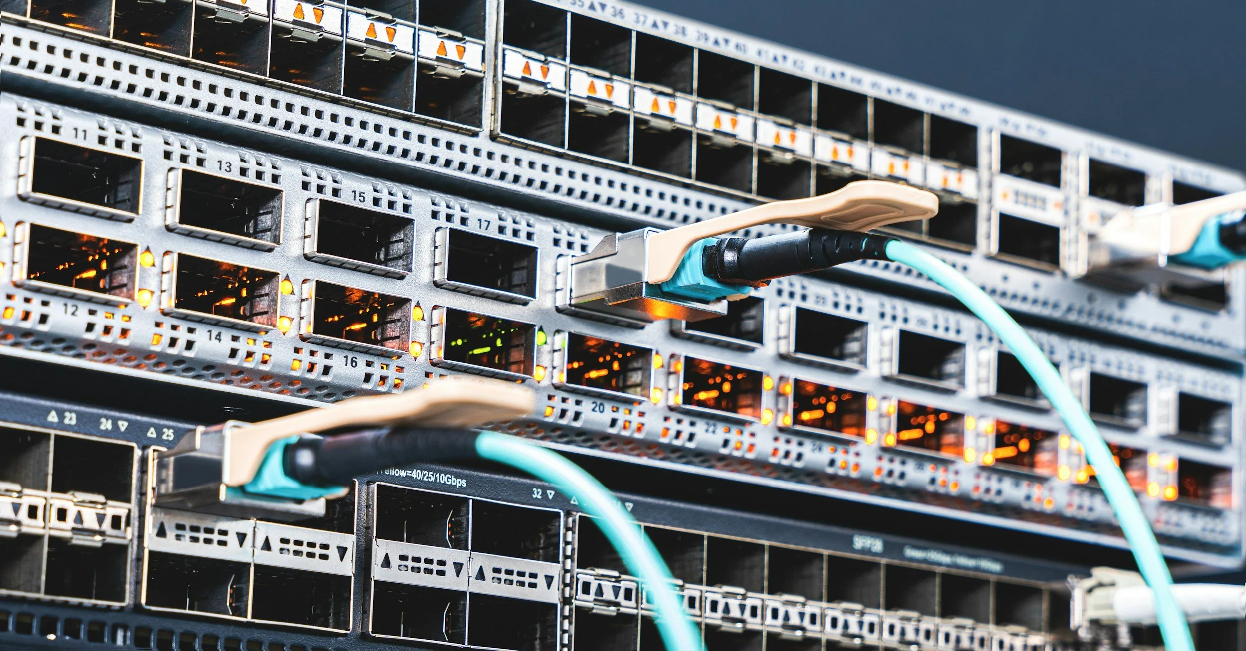 A young man with glasses and a beard, wearing a dark jacket, is working on wiring inside a modern server room or data center with numerous cables and illuminated indicator lights.