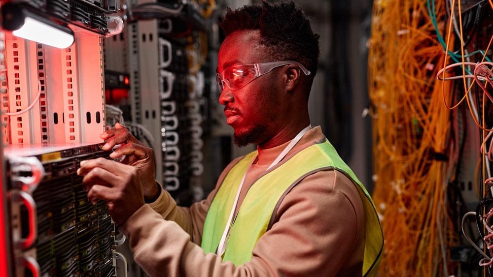 A technician wearing safety glasses and a high-visibility vest working on a server in a data center, surrounded by network cables and server equipment.