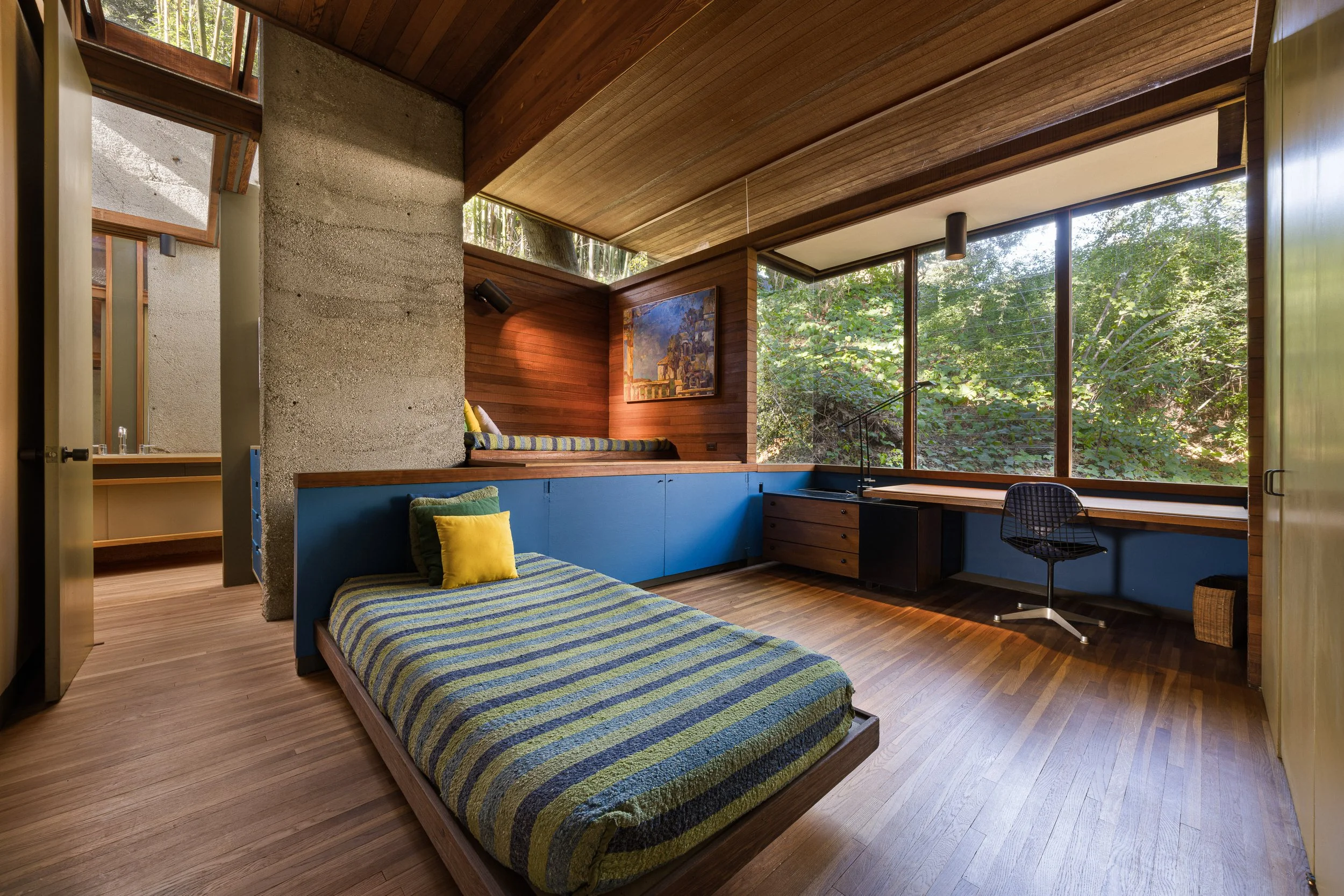 A bedroom with a single bed with a blue and green striped bedspread and pillows, large windows showing green foliage outside, wooden and concrete walls, a desk with a chair beside the window, and a built-in bench with a cushion and throw pillows.