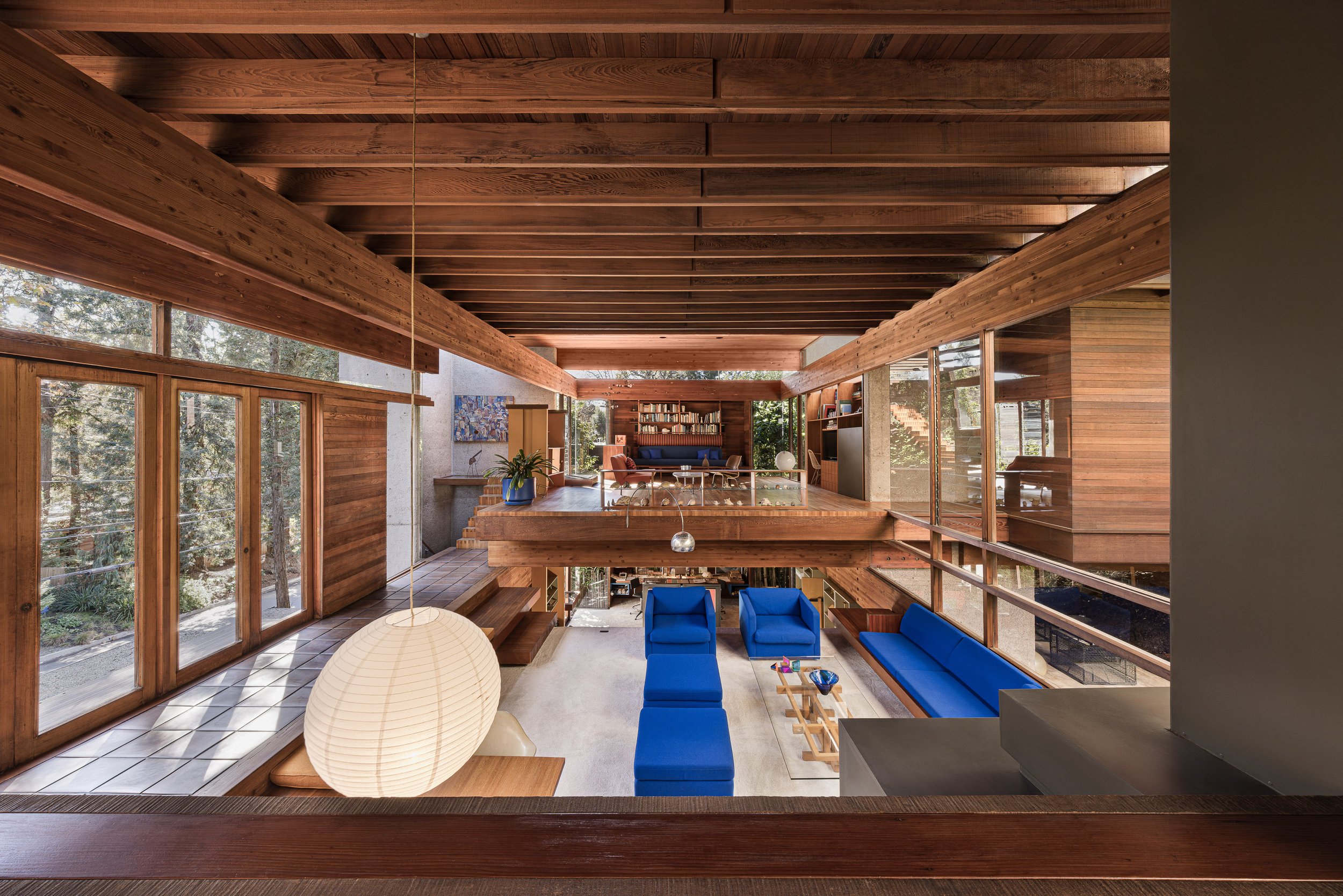 Interior view of a multi-level living room with extensive wooden paneling and large windows, featuring modern blue sofas and armchairs, a white paper lantern light fixture, and a bookshelf in the background.
