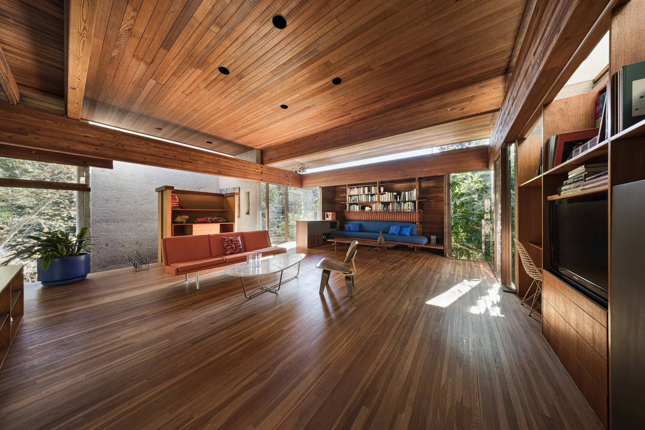 Living room with wooden flooring and ceiling, large windows with trees outside, orange sofa, wooden chair, glass coffee table, bookshelves, blue cushioned seating area, and various decorative items.